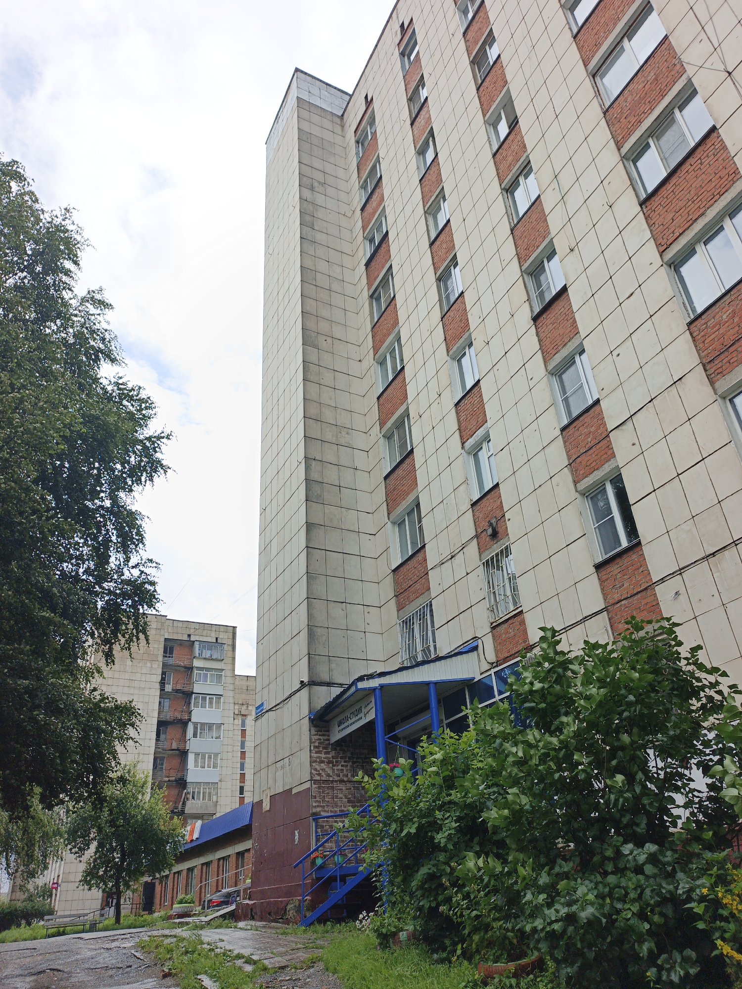 The image captures a multi-story apartment building in Zlatoust, Russia, under a cloudy sky. The building features a facade of beige tiles interspersed with red brick sections, and numerous windows. In the foreground, lush green trees and bushes partially obscure the lower floors and entrance of the building. A blue staircase leads to an entrance with a blue awning and a sign that reads "ШКОЛА-СТУДИЯ" (School-Studio). Further in the background, another apartment building is visible. The ground in front of the building is a mix of asphalt and overgrown grass.
