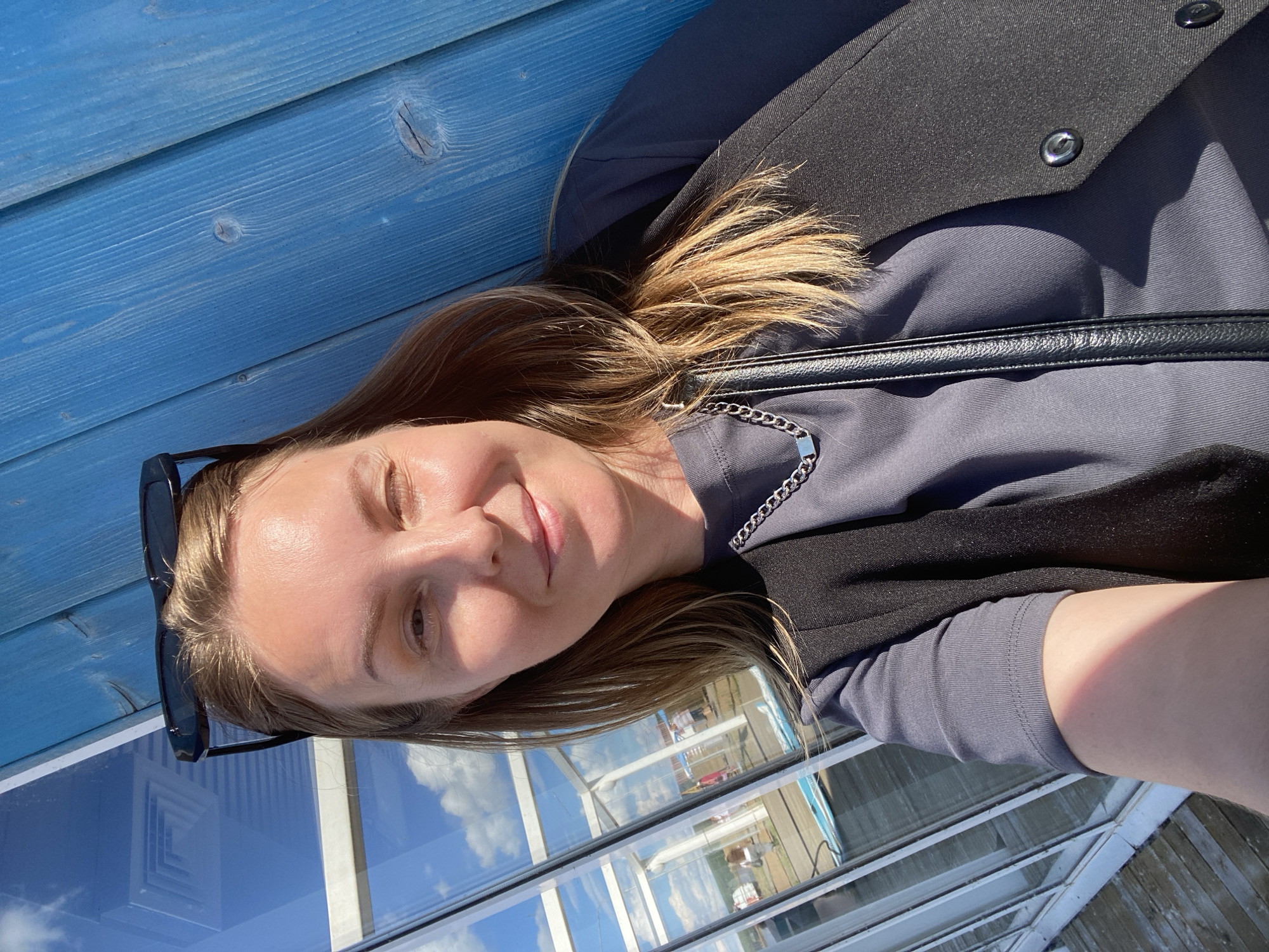 In a selfie taken outdoors, a woman with blonde hair and sunglasses perched on her head smiles as she leans against a bright blue wooden wall. She wears a dark grey t-shirt layered with a black vest and a silver chain necklace. To her left, a glass-paneled structure with white framing reflects the sky and clouds, revealing hints of people and equipment outside, possibly indicating an event or recreational area. The scene appears to be on a sunny day, with shadows casting across the woman's arm and clothing. The location is Chaikovsky, Russia.
