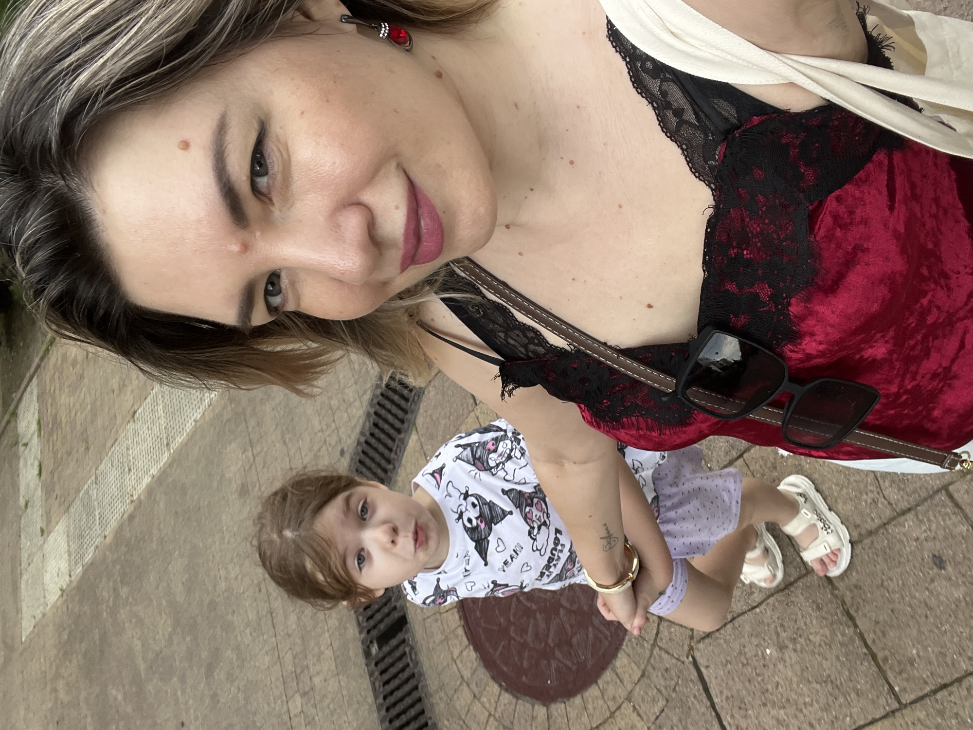 A woman and a young girl are standing outdoors on a paved walkway. The woman is holding the girl's left hand with her right hand. The woman is in the foreground, looking at the camera, while the girl is in the middle ground, looking slightly away from the camera with a surprised expression. The woman is wearing a red velvet camisole with black lace trim, a white bag over her right shoulder, and a red earring in her right ear. She has blonde hair with darker roots and visible moles on her face and neck. The girl is wearing a white t-shirt with a black and white cartoon print and light purple shorts. She has light brown hair and is wearing white sandals. In the background, there is a patterned paved surface and a drain grate. The context of Sochi, Russia, is not explicitly evident from the image itself.