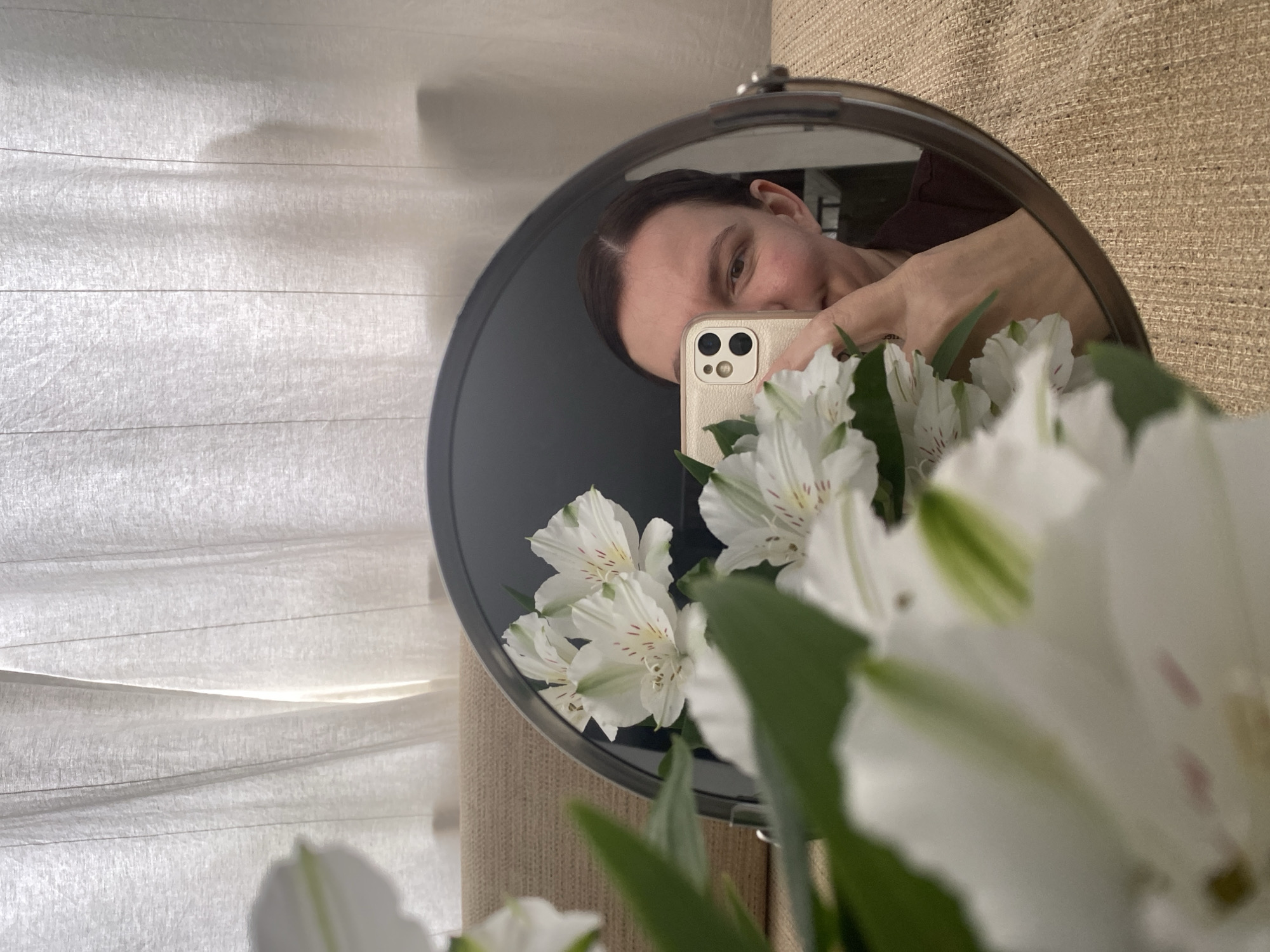 A selfie reflection in a round mirror shows a fair-skinned woman holding a tan-colored phone with a dual-lens camera. The woman's face is partially obscured by the phone. Her hair is dark and pulled back. The mirror also reflects a bouquet of white alstroemeria flowers with green leaves, partially obscuring the woman's reflection. In the background of the mirror are green trees and brown objects. The mirror is positioned in front of a beige fabric wall, and a semi-sheer white fabric panel is to the left of the mirror. This picture was taken indoors in Chaikovsky, Russia.