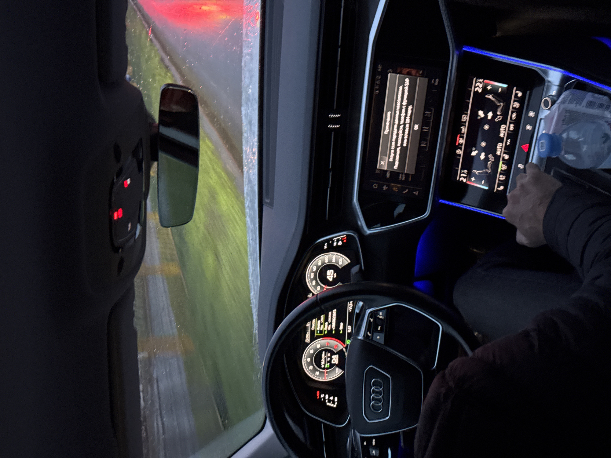 Here is a matter-of-fact description of the image:
The image shows the interior of an Audi car from the driver's perspective. The steering wheel with the Audi logo is prominently featured. The dashboard displays various digital information, including speed, navigation, and car settings. The infotainment screen shows a notification in Russian, which is related to network data services. The climate control panel is visible, with a hand near it, possibly adjusting the settings. A water bottle is present on the dashboard. Through the side mirror, there's a view of the outside environment, including the road and some greenery, possibly indicating the car is moving. The lighting is dim, with blue ambient lighting inside the car.