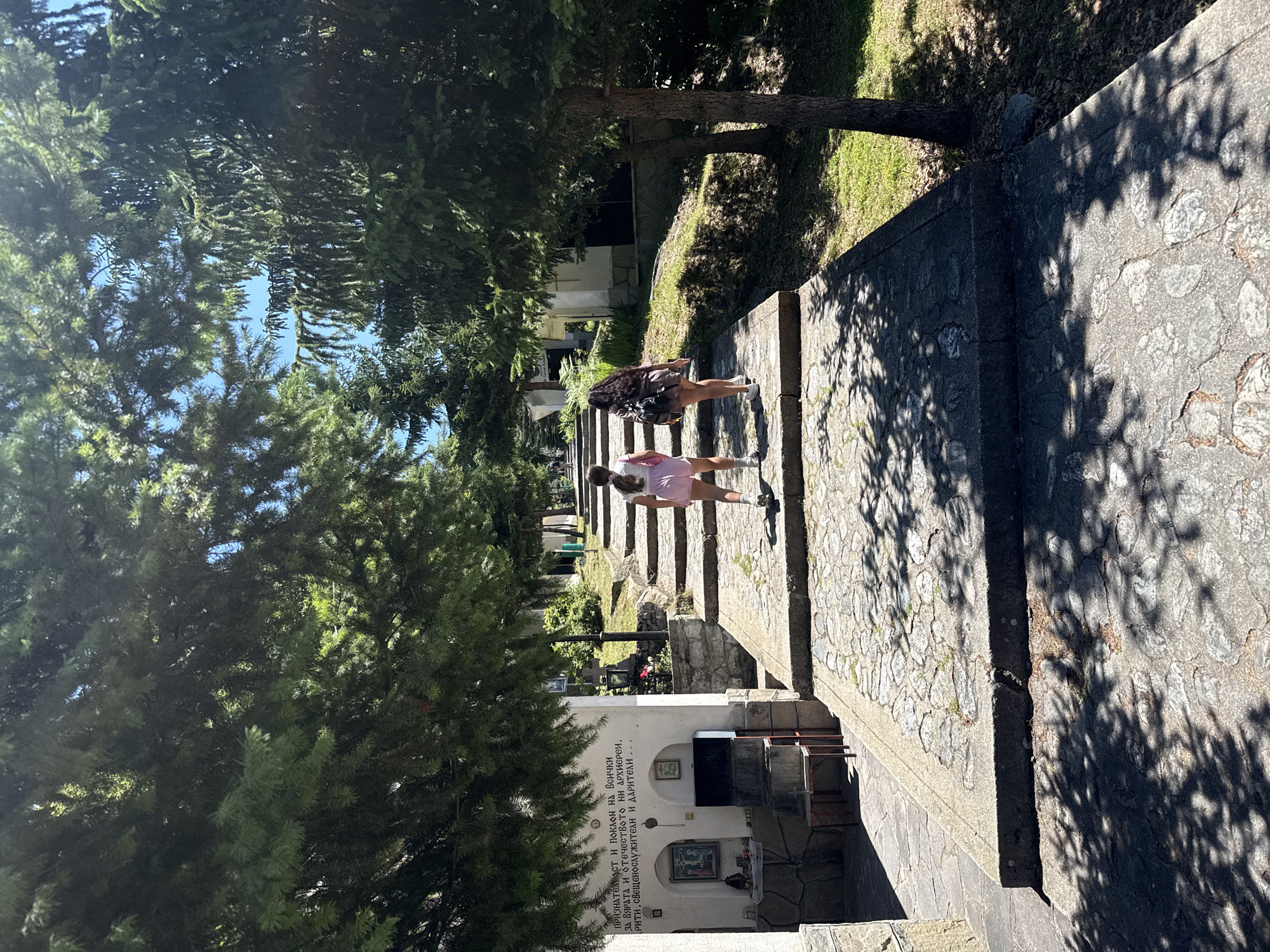The image shows two young females walking up a stone staircase lined with trees in Belitsa, Bulgaria. The staircase is made of irregular stone slabs with concrete edges. The trees are tall and their branches cast shadows on the staircase. To the left of the staircase, there is a white building with inscriptions and religious iconography. The building appears to be a small chapel or shrine. The sky is clear and blue. The females are walking away from the camera, one in a pink top and shorts, the other in a dark top and skirt. They are both wearing sandals. The environment appears to be a park or a sacred site.