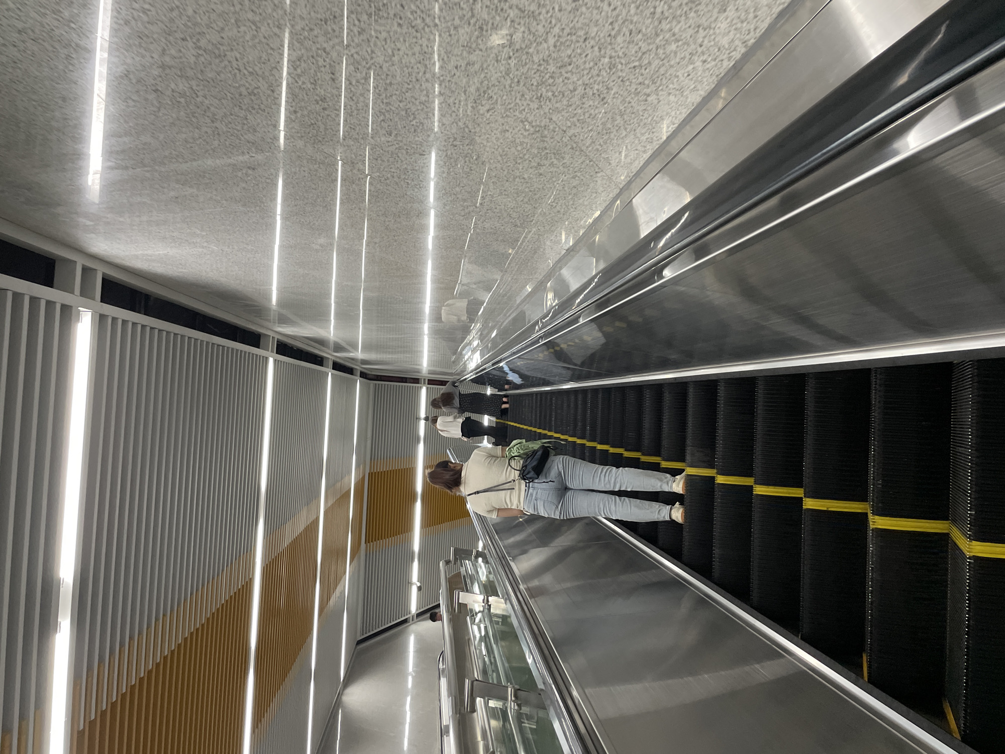The image depicts an escalator in what appears to be a modern transportation hub or public building, likely in the Liangjiang district of China, given the context. The escalator itself is black with yellow safety strips, ascending at a moderate incline. Several individuals are seen using the escalator. In the foreground, a woman with light-colored hair pulled back in a ponytail is walking up the escalator, wearing a light-colored short-sleeved shirt, light blue jeans, and white sneakers. She is carrying a dark purse with a green strap over her shoulder. Behind her, further up the escalator, two other figures are visible, one in a dark dress and the other in white attire.
The environment surrounding the escalator is characterized by sleek, contemporary design elements. The walls are clad in a speckled gray granite-like material, and the ceiling features a ribbed metal paneling system with integrated linear lighting. The handrails of the escalator are polished stainless steel. The overall impression is one of a clean, well-lit, and functional public space. The perspective is from behind and slightly below the main subject, looking upwards along the escalator's path.
