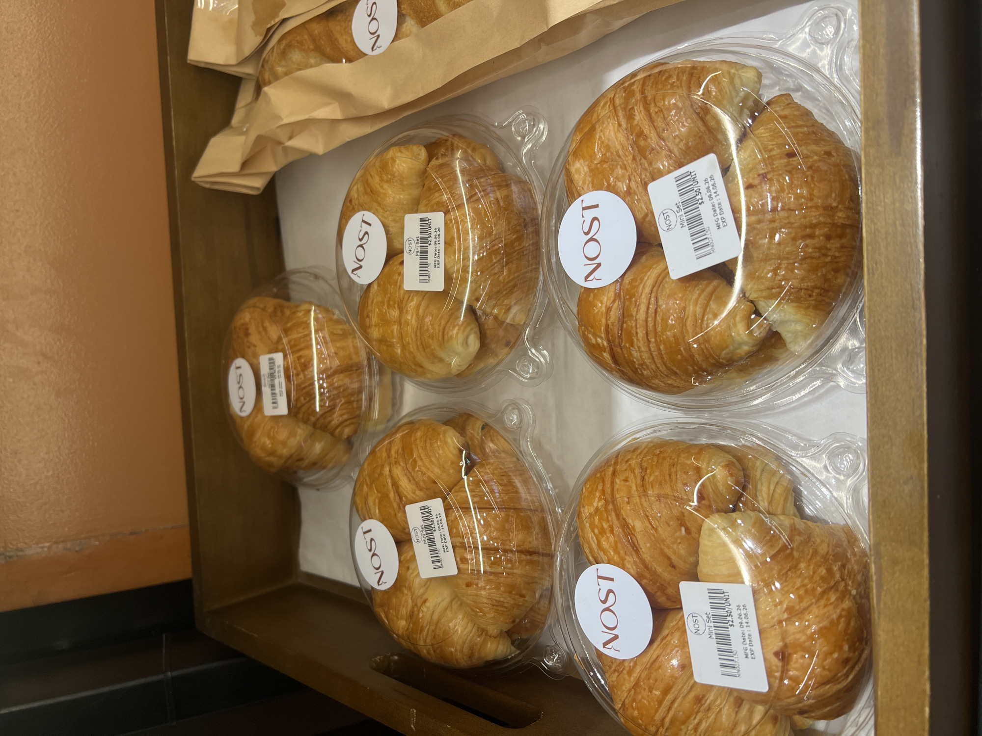 The image shows a close-up view of a wooden tray filled with individually packaged croissants. There are at least six packages of croissants visible, with most of them in clear plastic containers. Each container has a white circular sticker with the brand name "NOST" in red lettering. Some of the packages also have a white rectangular label with product information. The croissants appear golden brown and flaky. In the background, a brown paper bag containing croissants is partially visible. The overall impression is one of baked goods presented for sale. The context of Siem Reap, Cambodia is not directly evident from the visual cues in the image itself; it's a factual assertion provided in the prompt.
