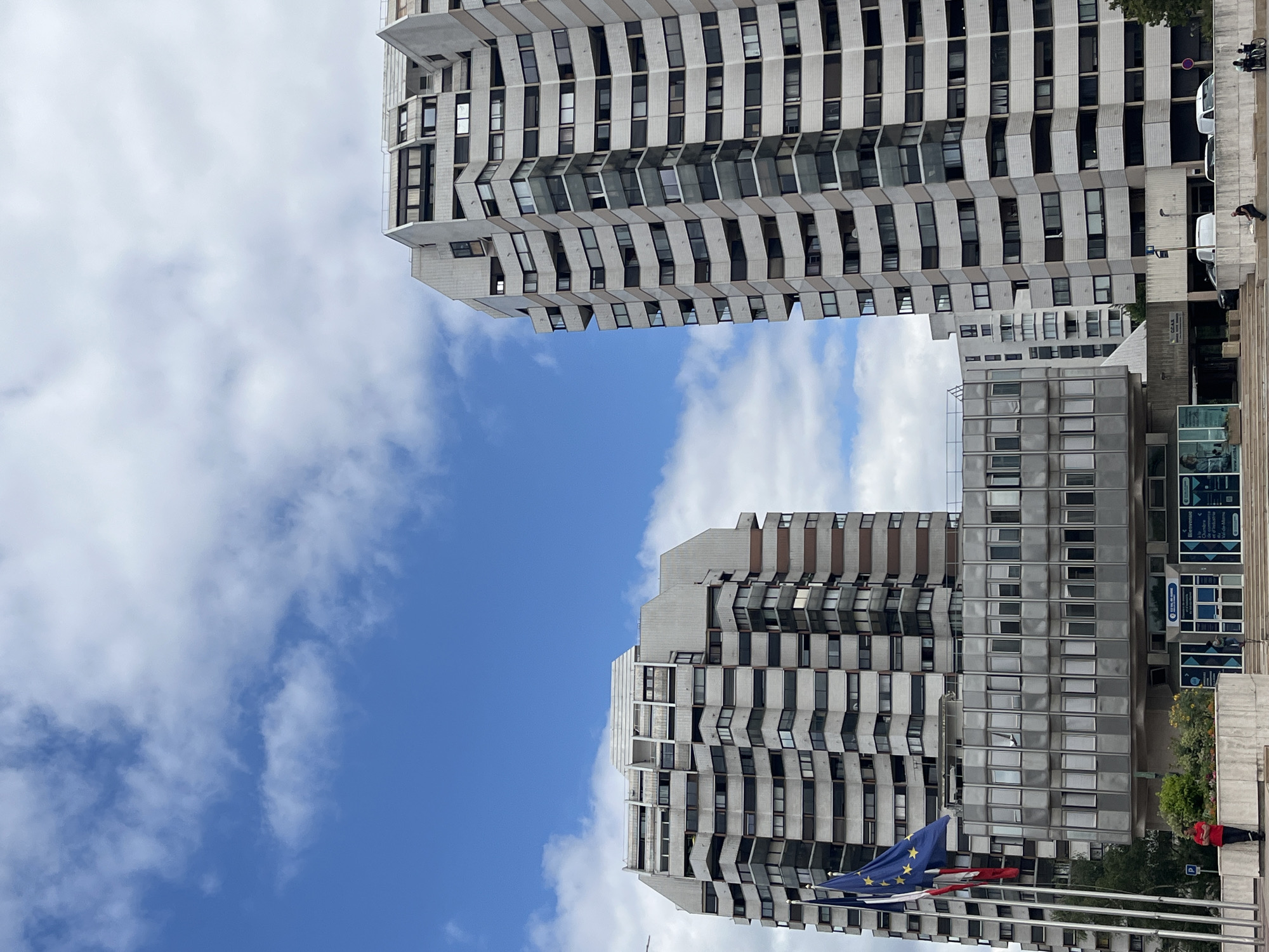 This media file shows a low-angle view of two large, modern apartment buildings against a bright blue sky with scattered white clouds. The buildings are tall and appear to be constructed from concrete and glass. Both have a repetitive, modular design with numerous windows. The building in the foreground has a grey facade and a distinctly zigzagging balcony structure. The building in the background, visible on the right, is also grey and has a more uniform facade with rows of windows.
In the lower right of the image, a few people are visible, including one person wearing a red jacket sitting on a ledge. There are also some parked vehicles, including what appears to be a white van. A flag, likely the European Union flag due to its blue color and yellow stars, is prominently displayed in the foreground, waving in the breeze. Other flags are also visible, suggesting a public space. Some signage is visible on the building in the background, but the text is too small to read clearly. The overall scene suggests daytime in an urban setting. The clear sky and bright lighting indicate fair weather. The architecture suggests a European location, consistent with the provided location context of Créteil, France.