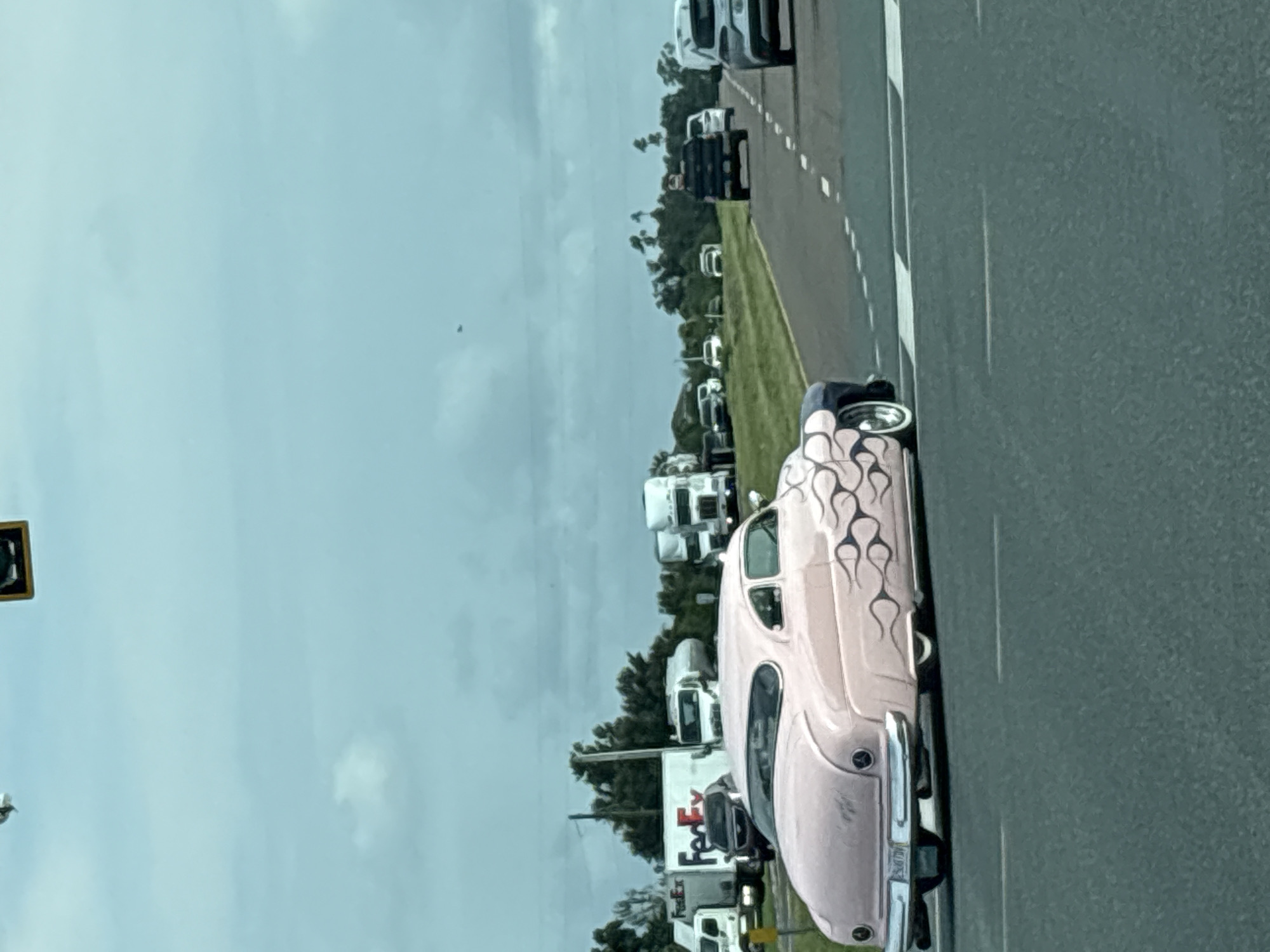 A pale pink classic car with black flame decals drives on a highway in Palmetto, United States. The car has a blue top and chrome accents. In the background, several FedEx trucks and other vehicles are visible. The sky is overcast with a few scattered clouds. The road appears to be a multi-lane highway.