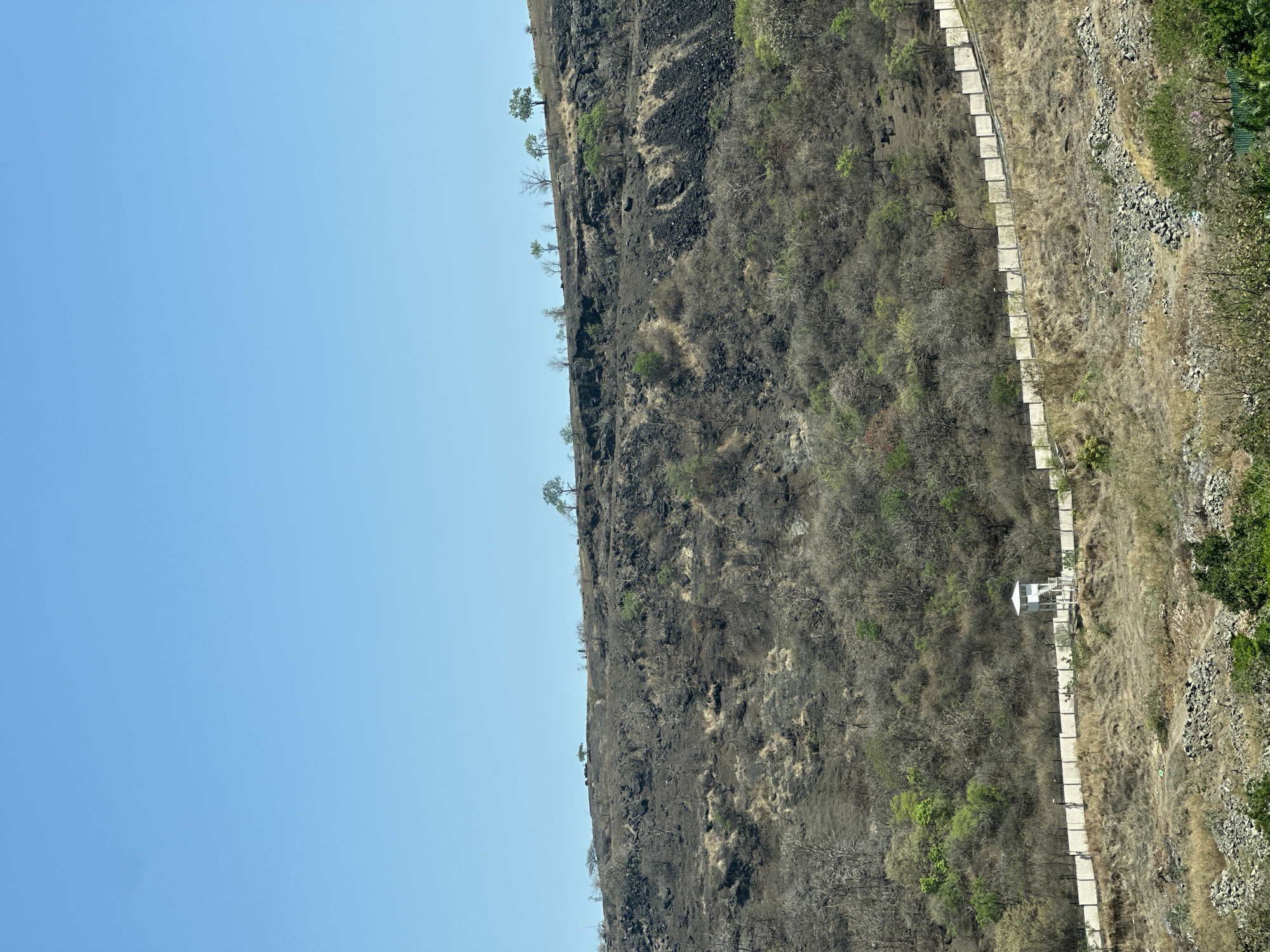 The image captures a steep, rocky hillside in what appears to be a semi-arid environment. A concrete walkway, lined with what looks like stone or concrete blocks, runs along the side of the hill. A small, white, gazebo-like structure is visible along the walkway. The hillside is covered with sparse vegetation, including dry grasses and some small trees. To the left of the hillside is a clear, blue sky. The location is likely near Man, India, given the prompt. The hillside seems to have signs of past burns, with patches of blackened earth.