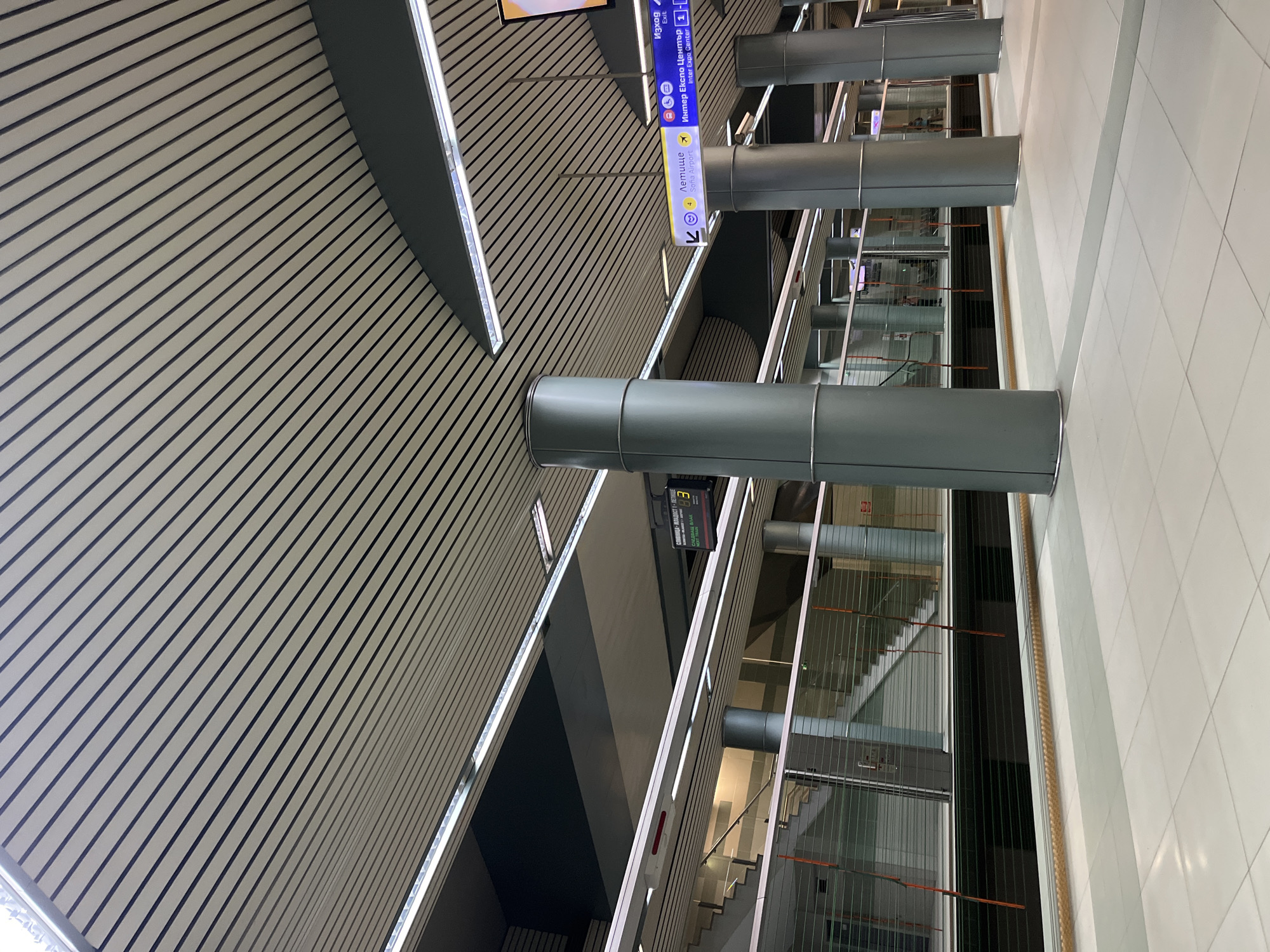 The image captures an interior view of what appears to be a modern architectural space, possibly within the Sofia Airport in Bulgaria, indicated by the airport signage in Bulgarian. The focus is on the building's infrastructure and design elements.
The left side of the frame features a wall with horizontal, linear patterns of light and dark shades, creating a striped effect. Integrated into this wall are sections of what appears to be recessed lighting. There are cylindrical support structures visible, running from the ceiling down. Signage indicating exits and directions to the airport and the Inter Expo Center is prominently displayed.
On the right side of the frame, a tiled floor suggests a walkway or public area. Further in the background, there's a multi-level structure with glass barriers, revealing staircases and possibly escalators. No people are visible. The scene is well-lit and oriented to emphasize the verticality and depth of the space.