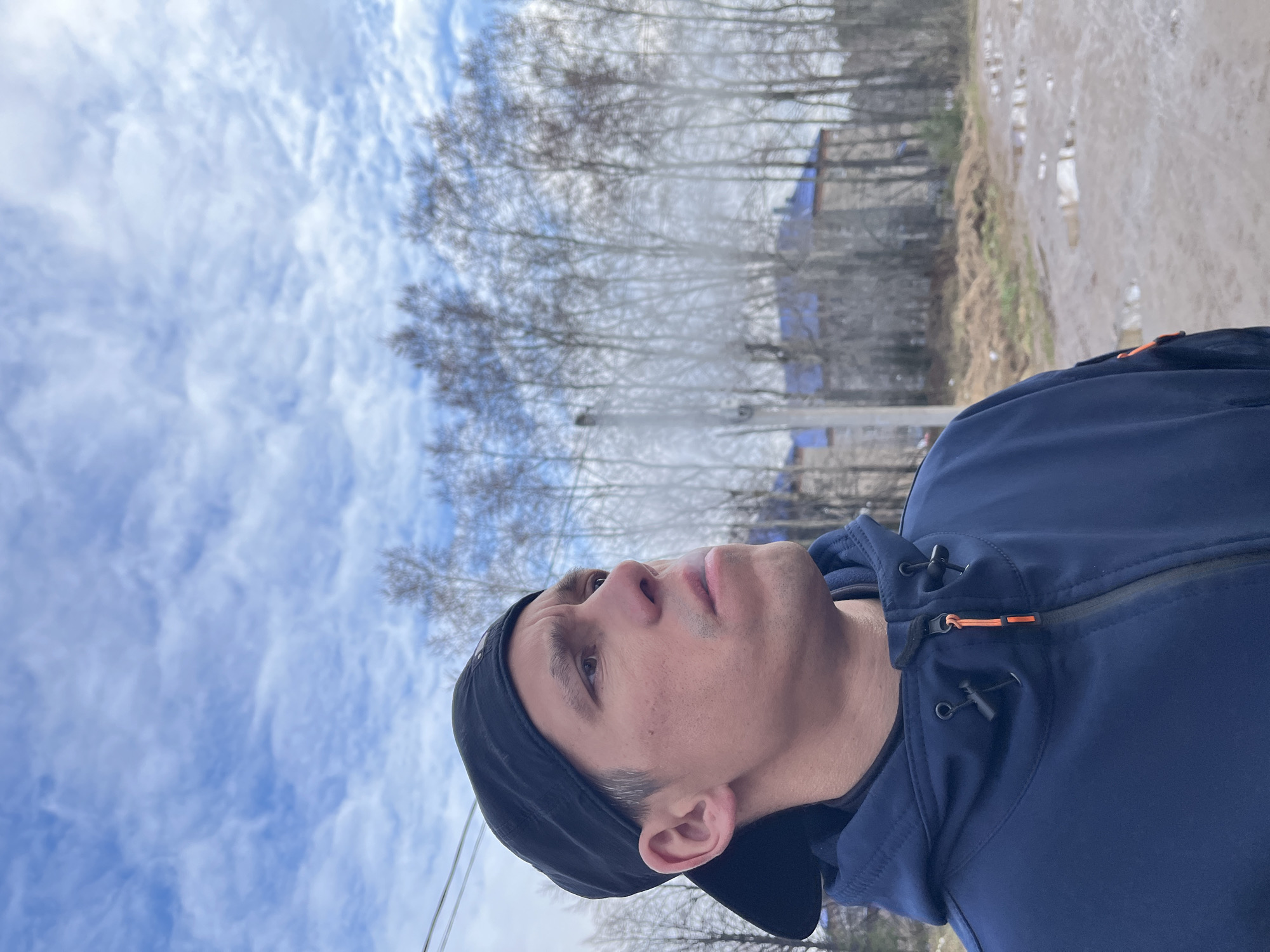 The image depicts a man wearing a dark blue jacket and a black cap looking upwards. Behind him is a view of a sky with scattered clouds, and a row of bare trees. A muddy path or area can be seen on the right side of the frame. Given the indication that the location is in Chaikovsky, Russia, it is plausible that the scene is a natural environment, possibly a park or outskirts of the city, photographed during the colder months when trees are leafless.