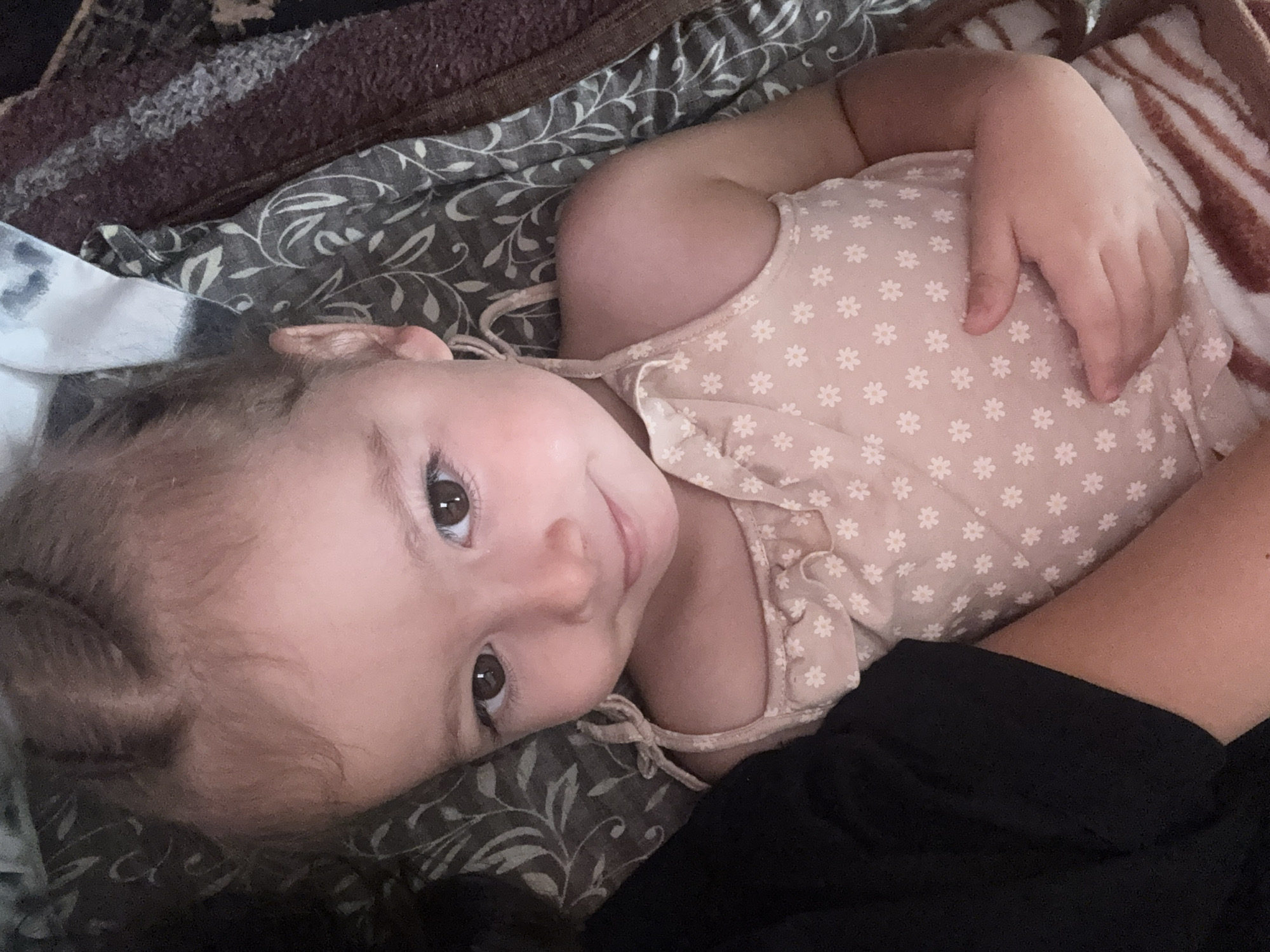 A close-up, eye-level shot focuses on a young child lying down, their head tilted slightly. The child has light brown hair styled in two braids, and dark brown eyes. They are wearing a light pink tank top adorned with small white floral patterns. Their right arm is bent, with their hand resting on their stomach. The child's left side is partially obscured by a black garment. The background consists of patterned fabric, appearing to be a blanket or bedding, with shades of gray, white, and brown. The image was taken indoors in Basarabeasca, Moldova.