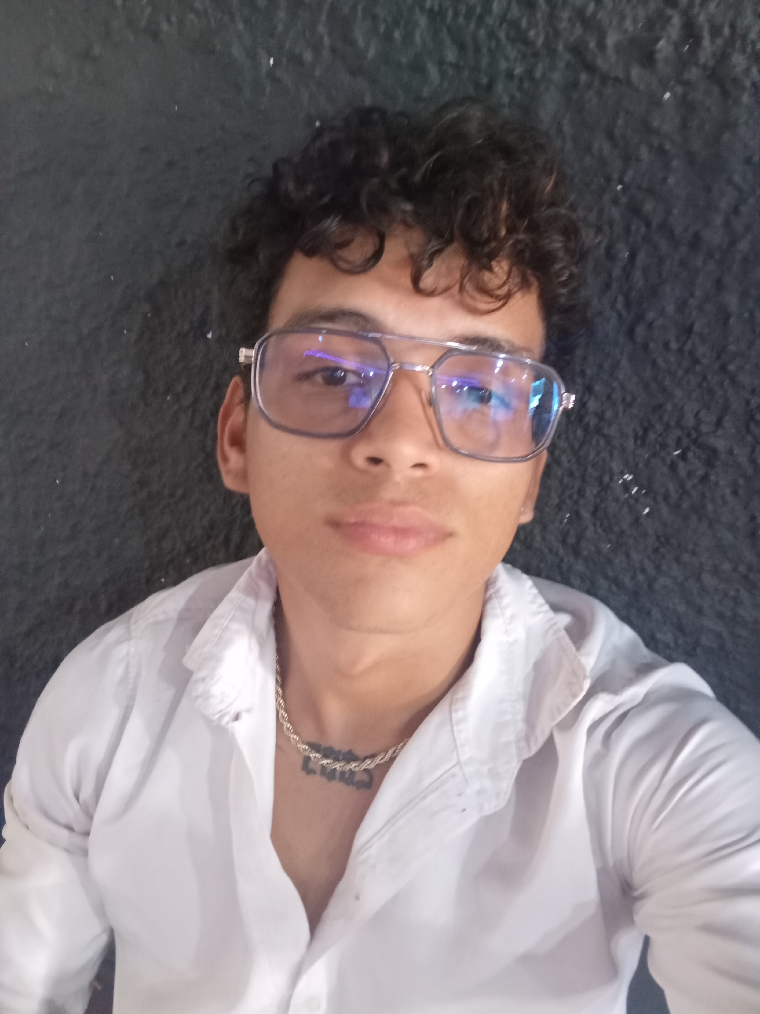 A young man with curly dark hair and glasses is wearing a white collared shirt and a gold chain with a tattoo visible underneath. He is looking at the camera, and his face is well-lit. The background is a dark, textured surface, possibly a wall or fabric. The image appears to be a selfie taken indoors. There are no discernable elements to suggest a specific location in Juan Griego, Venezuela.