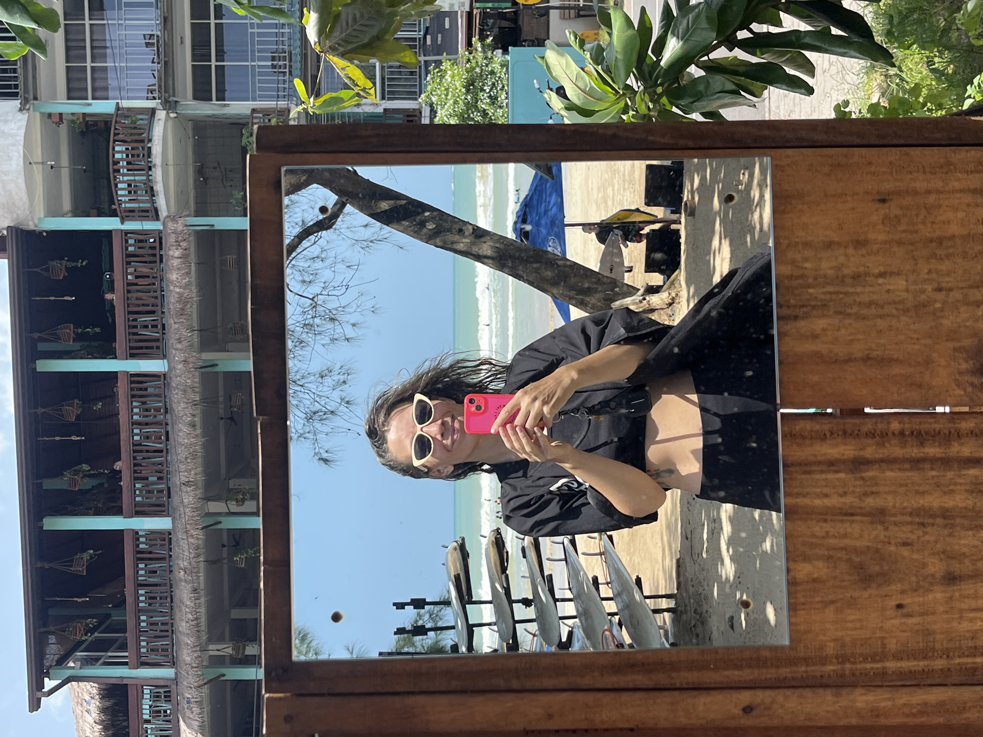 A woman takes a selfie in a mirror at the beach in Weligama, Sri Lanka. The woman is smiling and holding a pink smartphone. She is wearing a black crop top, black pants, and white sunglasses. In the background, there are surfboards, the ocean, and a building with multiple levels and balconies. A tree is visible in the mirror, and green leaves are in the foreground on the right side of the image. The scene appears to be a sunny day.