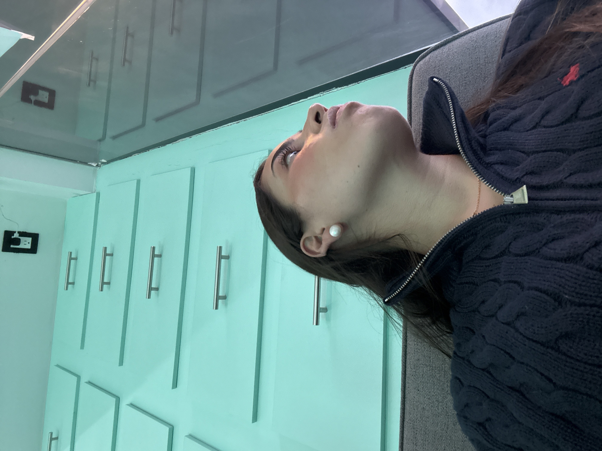A woman is reclining in a gray chair, looking up and to the left. She is wearing a navy blue cable knit sweater with a partial zipper. Her skin appears fair and she is wearing a pearl earring. Her dark hair is visible near her ear and at the top of the frame.
The background consists of mint green cabinets with silver handles. A gray ceiling is visible above the cabinets. A black electrical outlet is seen on the wall next to the cabinets.
The scene appears to be indoors. I do not have enough information to confirm the city of San Bernardino Tlaxcalancingo, Mexico, as the location.