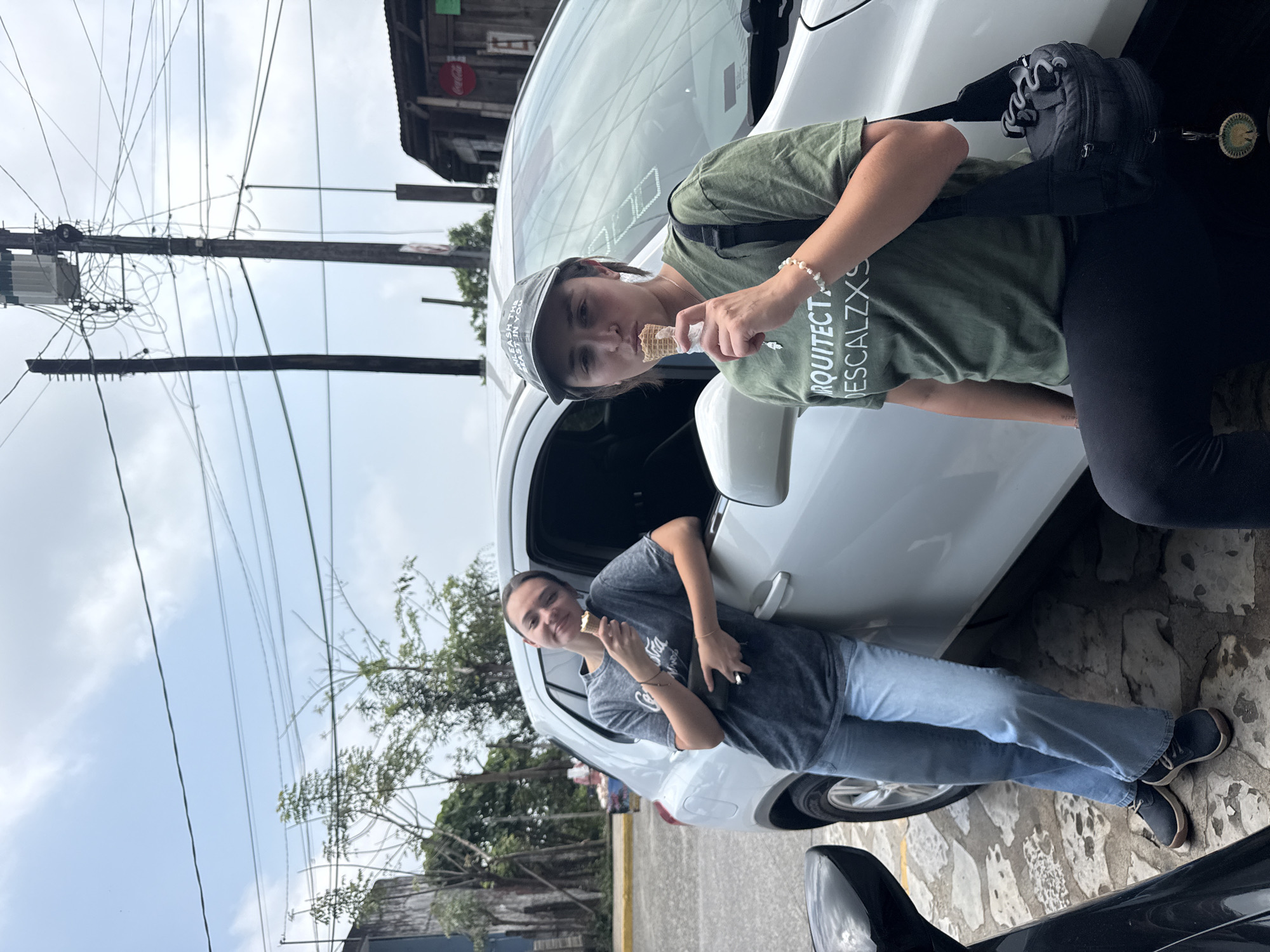 Two young women are standing near a white car, both holding and appearing to be eating ice cream cones. The woman on the left is wearing a gray t-shirt and light blue jeans. The woman on the right is wearing an olive green t-shirt and black pants. In the background, there are buildings and power lines, suggesting an urban setting. The location is Cuetzalan, Mexico.
