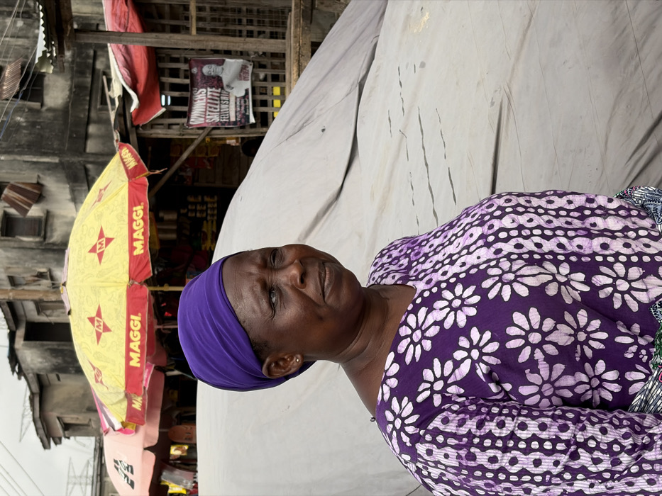 The image depicts a woman standing in an outdoor market setting within Jakande, Nigeria, under an overcast sky. She is positioned slightly off-center to the left, facing towards the right with a neutral expression. The woman has dark skin and wears a vibrant purple headwrap, a purple V-neck top with a white floral pattern, and the edge of a multi-colored patterned fabric is visible at her waist. Her left eye (viewer's right) exhibits a noticeable bluish tint. In the background, a bustling market environment is visible with several stalls and structures. A prominent yellow umbrella bearing multiple "MAGGI." logos in red provides shade over a stall displaying various packaged goods. To the left, a partial pink umbrella is visible. Behind these, distressed multi-story concrete buildings with visible wear, some featuring corrugated metal elements, form the urban backdrop. A large white tarpaulin-covered object occupies much of the middle ground behind the woman. To the far right, a poster is affixed to a wooden structure, partially obscured, with the legible text "WINNERS' SATELLITE FELLOWSHIP" and an image of a man. Utility lines are discernible in the upper left corner of the background.