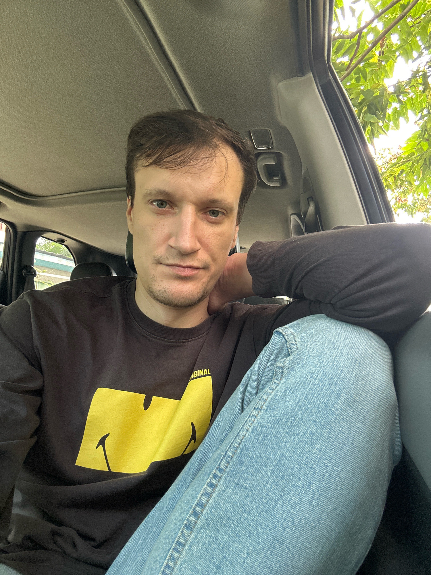 The image shows a young man sitting in the passenger seat of a car, with a selfie-style perspective. He is wearing a black sweatshirt with a large yellow graphic of a smiley face on it, and light-wash blue jeans. His right arm is resting on his left arm, which is bent at the elbow and rests on his leg. The man has light skin, short brown hair, and blue eyes. The interior of the car is visible, including the headliner, a sun visor, and a side door panel. Outside the car, through the open window, green foliage and the sky are visible. The scene appears to be outdoors, possibly in a residential area or park. There is no visible indication of the location being in the city of Makiv, Ukraine.