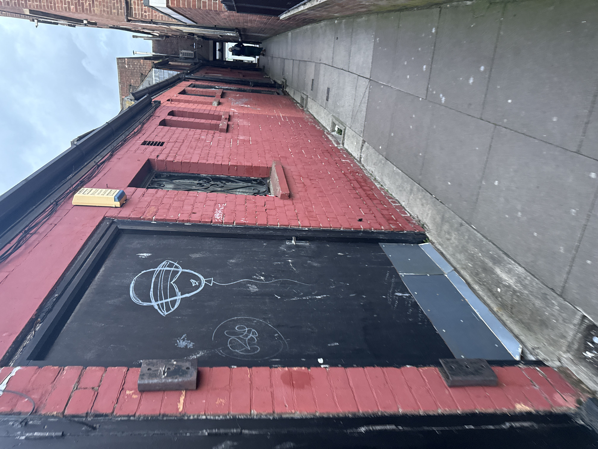 The image depicts a narrow alleyway in Great Yarmouth, United Kingdom. A red brick building lines the left side of the alley, its lower portion covered with a black painted section featuring graffiti, including a drawing of a balloon. Above the graffiti is a window with ornate metalwork. A yellow box is affixed to the brick wall above the black painted section. The alleyway floor is paved with gray concrete slabs, and a concrete wall runs along the right side. A person is faintly visible in the distance at the end of the alley. The sky is overcast.