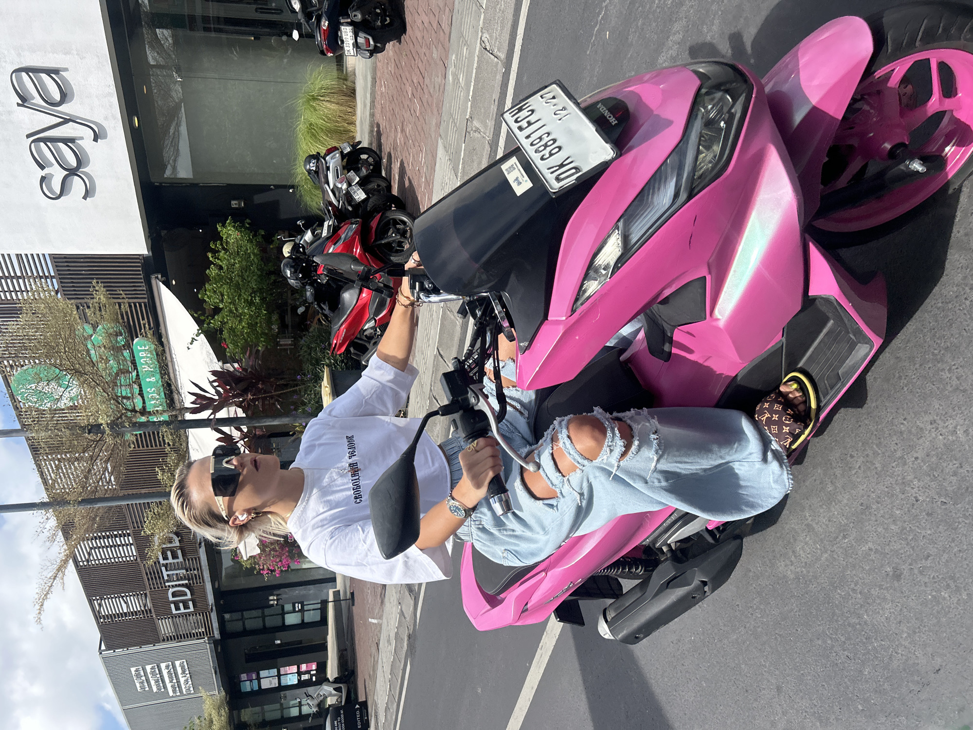 A young Caucasian woman with blonde hair pulled back and large black sunglasses is riding a bright pink scooter down a street in North Kuta, Indonesia. She is wearing a white cropped t-shirt with text in Cyrillic script and distressed light-wash denim jeans. Her left hand is on the scooter's handlebar, and her right hand is resting on her lap. She is wearing a wristwatch and slides on her feet. The scooter has a black windshield with a license plate that reads "DK 6891 FCH". In the background, there are several buildings with signage including "saya" and "EDITED". Other parked scooters and motorcycles are also visible. The street appears to be paved, and there is a sidewalk lined with bricks. The sky is bright and sunny.