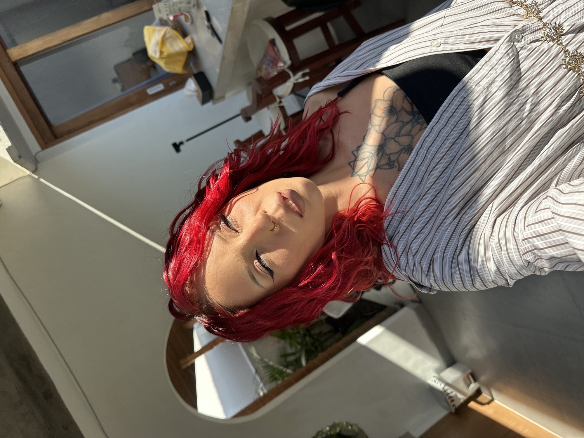 A close-up portrait shot captures a person with vibrant red, wavy hair, angled slightly upwards, possibly in a selfie. The individual has light skin, winged eyeliner, and a small nose ring. They are wearing a light-colored, vertically striped button-up shirt, unbuttoned to reveal a black top or bra underneath. A prominent black ink tattoo, depicting floral or geometric patterns, is visible on their left shoulder and upper chest. A gold chain with decorative sun-like medallions adorns the shirt.
The strong, natural sunlight casts defined shadows across the scene and illuminates the person's face and hair, suggesting it is daytime, likely morning or late afternoon. The background is softly blurred but shows elements of an indoor setting, possibly a room with white walls. Visible details include a wooden-framed window reflecting bright light, hinting at an outdoor view, and what appears to be a table or counter with a yellow cloth and other indistinct items. A chair is partially visible in the mid-ground. The overall impression is one of warmth and brightness. The person's expression appears relaxed, with eyes partially closed, giving a serene or contemplative mood. No visible text is present in the image. The scene is set in North Kuta, Indonesia.