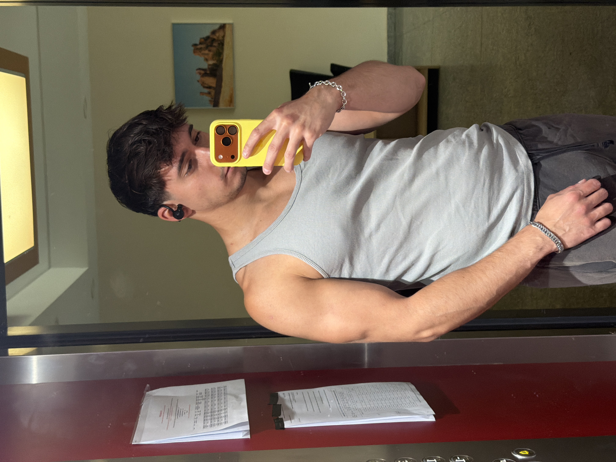 A young male, appearing to be in his early twenties, is captured in a mirror selfie within an indoor setting, likely an elevator lobby or hallway in Sofia, Bulgaria. He is looking down at his bright yellow smartphone, which he holds with his right hand to take the photograph. He wears a light grey ribbed tank top and dark grey sweatpants or joggers. A black wireless earbud is visible in his left ear, and silver chain link bracelets adorn both wrists. His hair is dark and styled with some texture, and he has light facial stubble.
The background, visible in the mirror's reflection, includes a prominent red wall panel on the left, to which several white documents with text and tabular data are affixed with clips or tape. Adjacent to this red panel is a darker vertical strip featuring silver elevator call buttons, one of which (marked '4') is illuminated in yellow, confirming the location. Further back in the reflection, behind the individual, there is a light beige wall with a rectangular ceiling light fixture that has a golden frame. A small landscape painting depicting arid, rocky terrain under a light blue sky hangs on this wall. A glimpse of dark furniture is visible in the far background, and the floor appears dark and somewhat reflective.