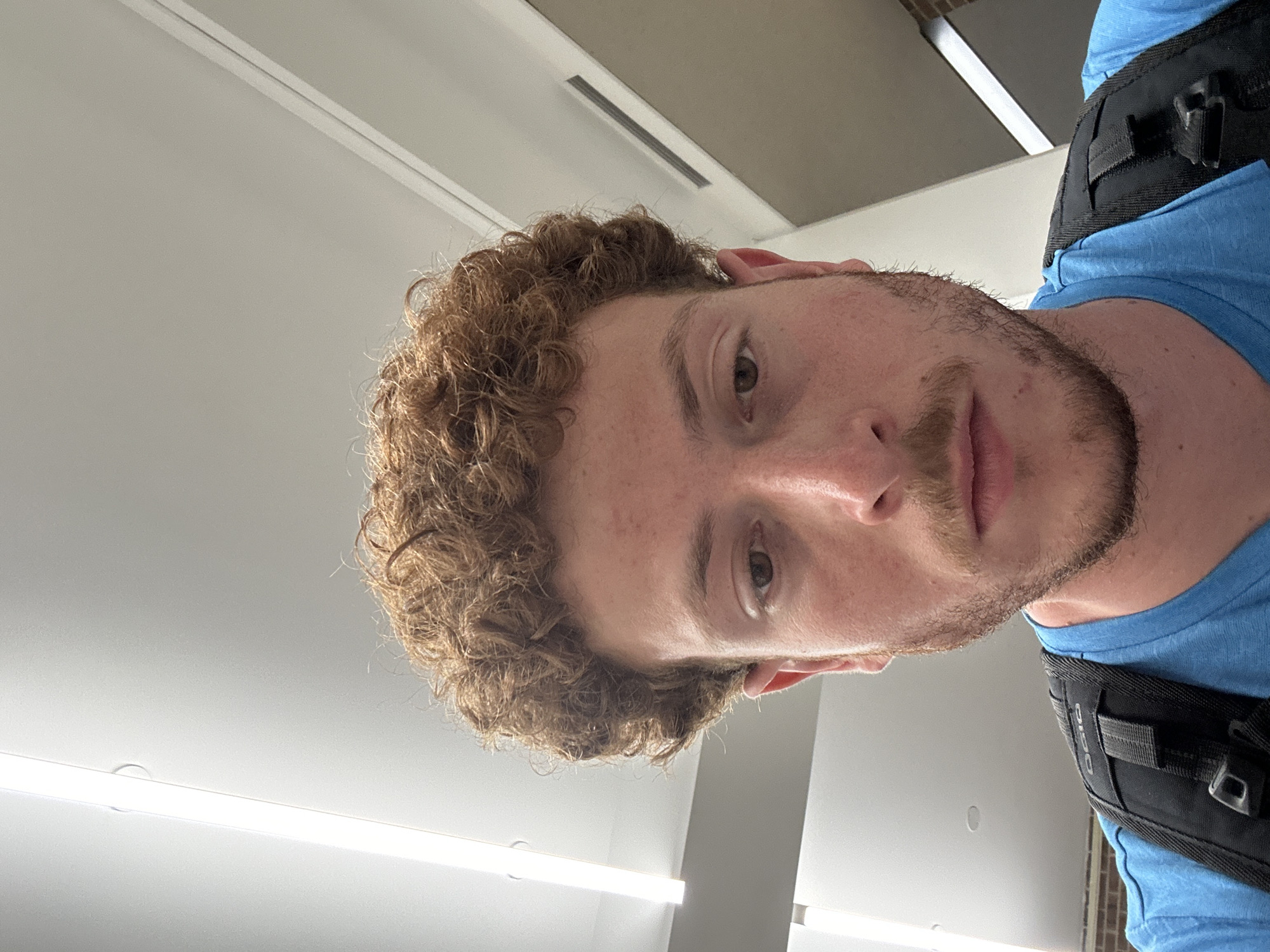 A close-up, selfie-style photo features a light-skinned individual with curly brown hair and a beard. The person is wearing a blue shirt and a black harness-like strap is visible over their shoulder. The background consists of a white ceiling with recessed lighting fixtures and air vents. There is insufficient information to determine the location within New York City.