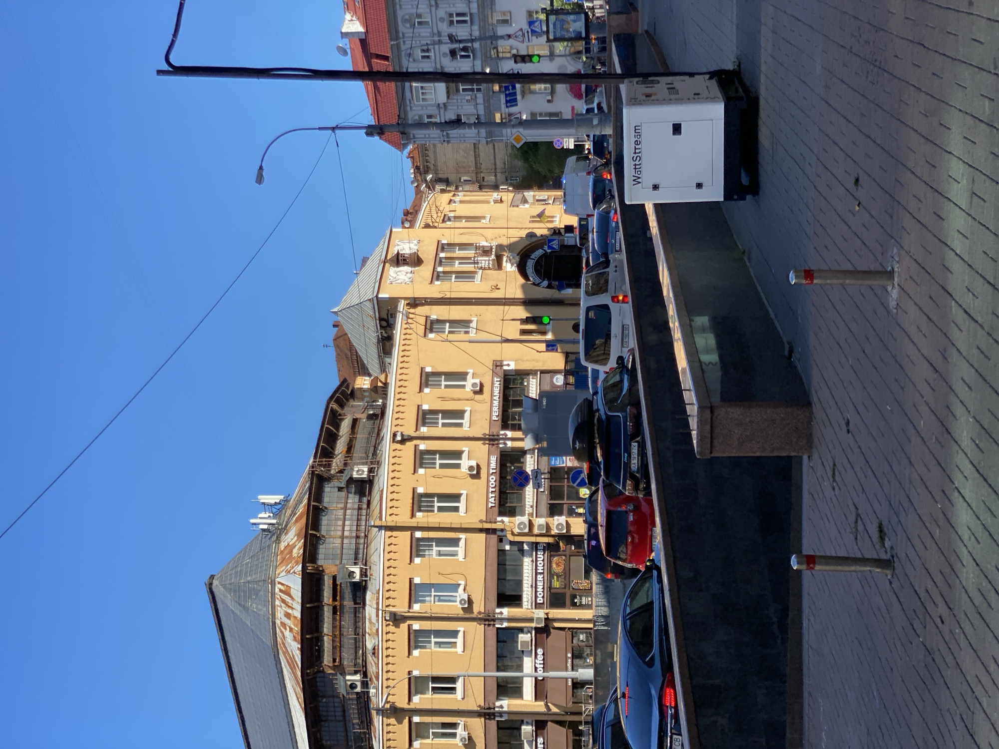 The image shows a street scene in a city under a clear blue sky. In the foreground, a paved walkway with bollards lines the right side. A white portable generator with "WattStream" branding sits on the walkway. A dark stone wall runs along the left side of the walkway, behind which is a street with several cars. The cars are stopped in traffic, and some are in lanes that appear to be descending. Buildings line the street, with a prominent yellow building on the left featuring several shops, including "Coffee," "Doner House," "Tattoo Time," and "Permanent." This building has a distinctive rounded, multi-story facade with a rusty, domed roof structure above. Across the street, other buildings of varying architectural styles are visible. Traffic lights are present and appear to be green for traffic moving from left to right. The overall impression is of a busy urban environment on a sunny day.