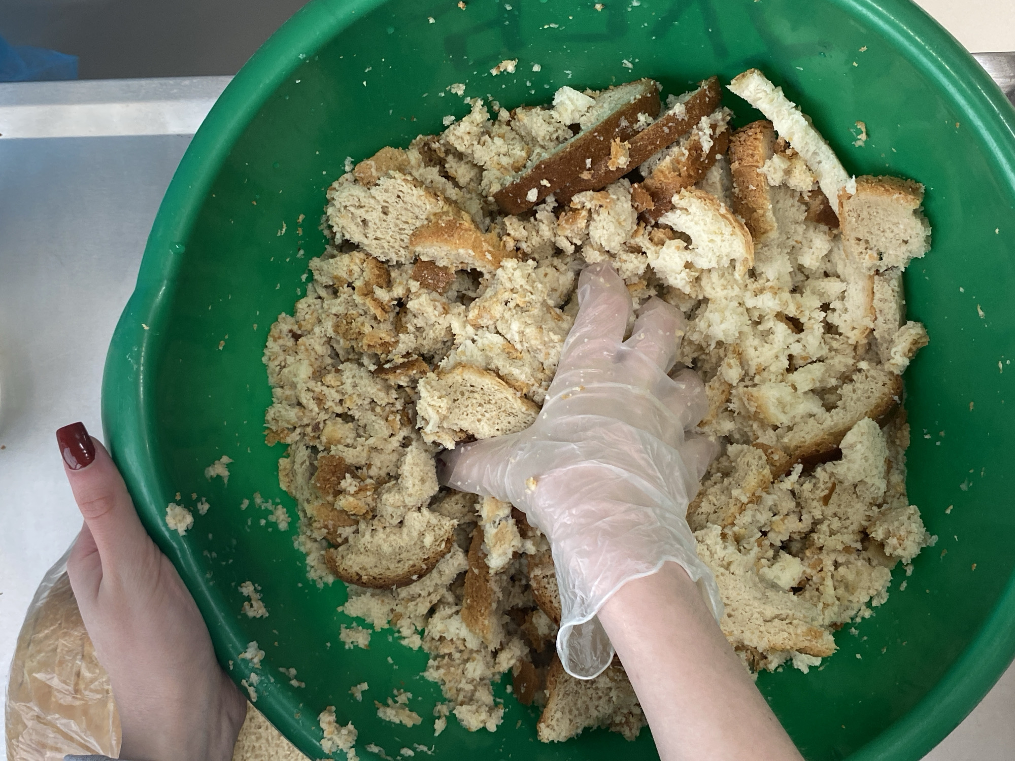 A top-down shot shows a person's gloved hand in a green plastic bowl filled with bread pieces in Марковский, Russia. The bread appears to be a mixture of white and brown varieties, cut into irregular sizes. The hand is covered in a clear plastic glove, suggesting food preparation. Another hand with dark red nail polish is visible on the left side of the frame, holding the bowl. The background appears to be a countertop.