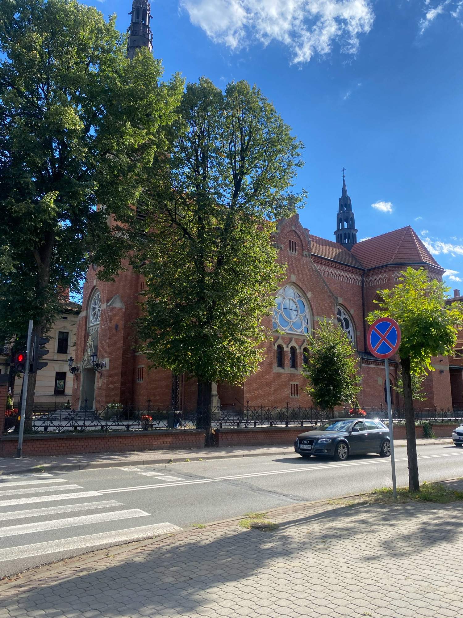 This image shows a street scene in Nowy Sącz, Poland, with a large brick church as the dominant background element. In the foreground, a pedestrian crossing is clearly marked on an asphalt road. A grey Audi car is traveling on the road, heading towards the right side of the frame. To the right of the car, a "No Stopping" sign is visible. Tall, leafy green trees partially obscure the church and the sky. The church itself is constructed of red brick and features a tall, dark spire with a cross at its apex. It has arched windows, including a prominent rose window. The sky is blue with scattered white clouds. The scene appears to be set on a sunny day.