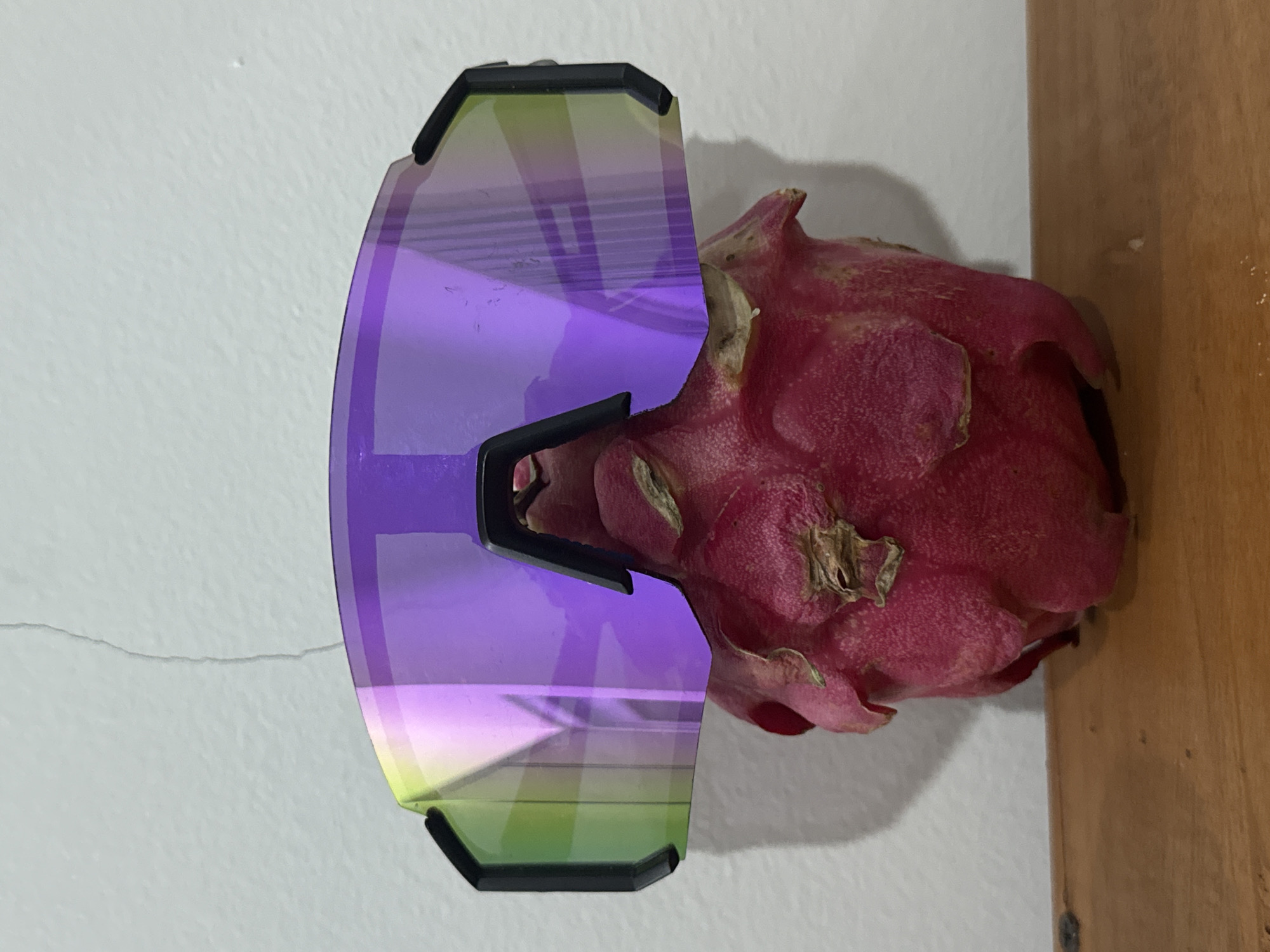 On a light grey wall with a horizontal crack, lies a pair of purple and green-tinted sports sunglasses with black frames. To the right of the glasses is a whole, unpeeled dragon fruit. The setting implies an indoor environment, but no details suggest a specific location in Kuta, Indonesia. No people or actions are visible.