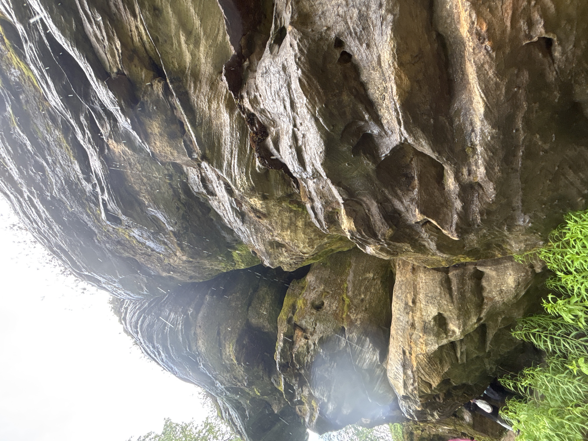 The image captures a low-angle view of a large rock formation, possibly a cliff face, on a cloudy day. Water is visible falling in streaks down the rock, suggesting rain or a nearby waterfall. The rock is layered and textured, with patches of moss and evidence of erosion. In the lower left portion of the image, a few individuals are partially visible, appearing to be hiking or exploring near the base of the rock formation. One person is wearing a pink jacket, and another has a white backpack. Lush green vegetation, likely tall grass or small plants, grows at the very bottom of the frame. The overall environment appears natural and outdoors. There is no information available that places this specific scene within the city of Shumikhinskii, Russia.