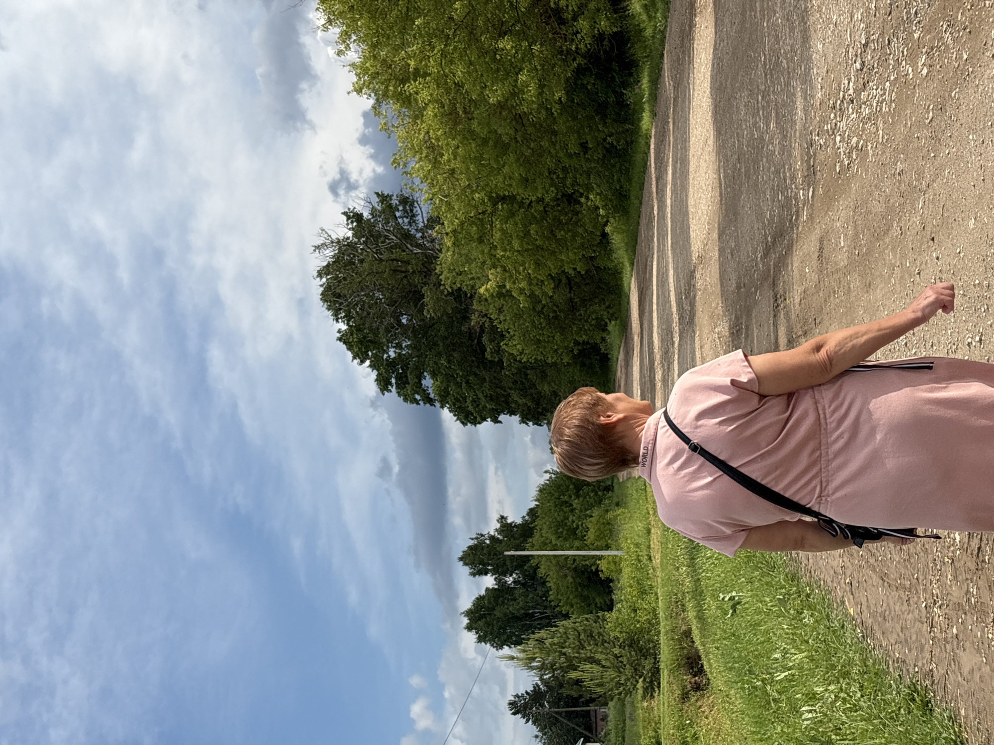 A woman wearing a pink dress and a black crossbody bag walks on a dirt road in a rural area. She is facing away from the camera, looking down the road. The road is flanked by green grass and trees on either side, with a bright blue sky and some clouds overhead. The scene suggests a warm, sunny day in a natural setting. The location is identified as Kumyonya, Russia.