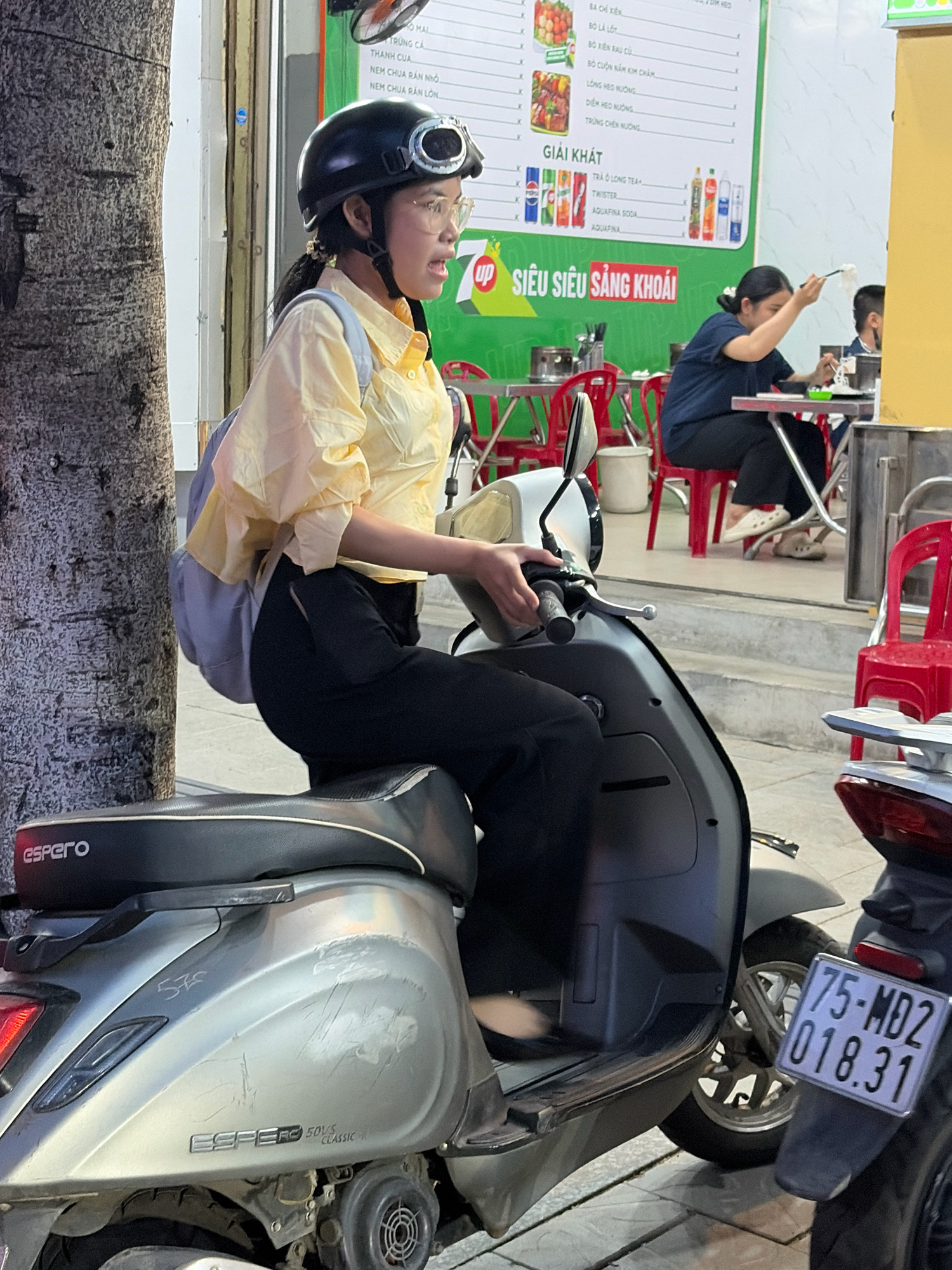 The image shows a young woman wearing a black helmet and glasses, sitting on a silver scooter. She is wearing a yellow shirt and black pants, and has a light blue backpack on. She appears to be talking and looking to her right. The scooter has "ESPERO" written on the seat and "ESPERO 50S CLASSIC" on the side. To the right of the scooter, another scooter is visible with a license plate that reads "75-MD2 018.31". In the background, there is a restaurant with red chairs and a menu board displaying Vietnamese text and images of food and drinks. This scene is likely located in the city of Đà Nẵng, Vietnam, as indicated by the license plate prefix "75" which corresponds to Đà Nẵng. The overall environment appears to be an outdoor dining area of a street-side restaurant.