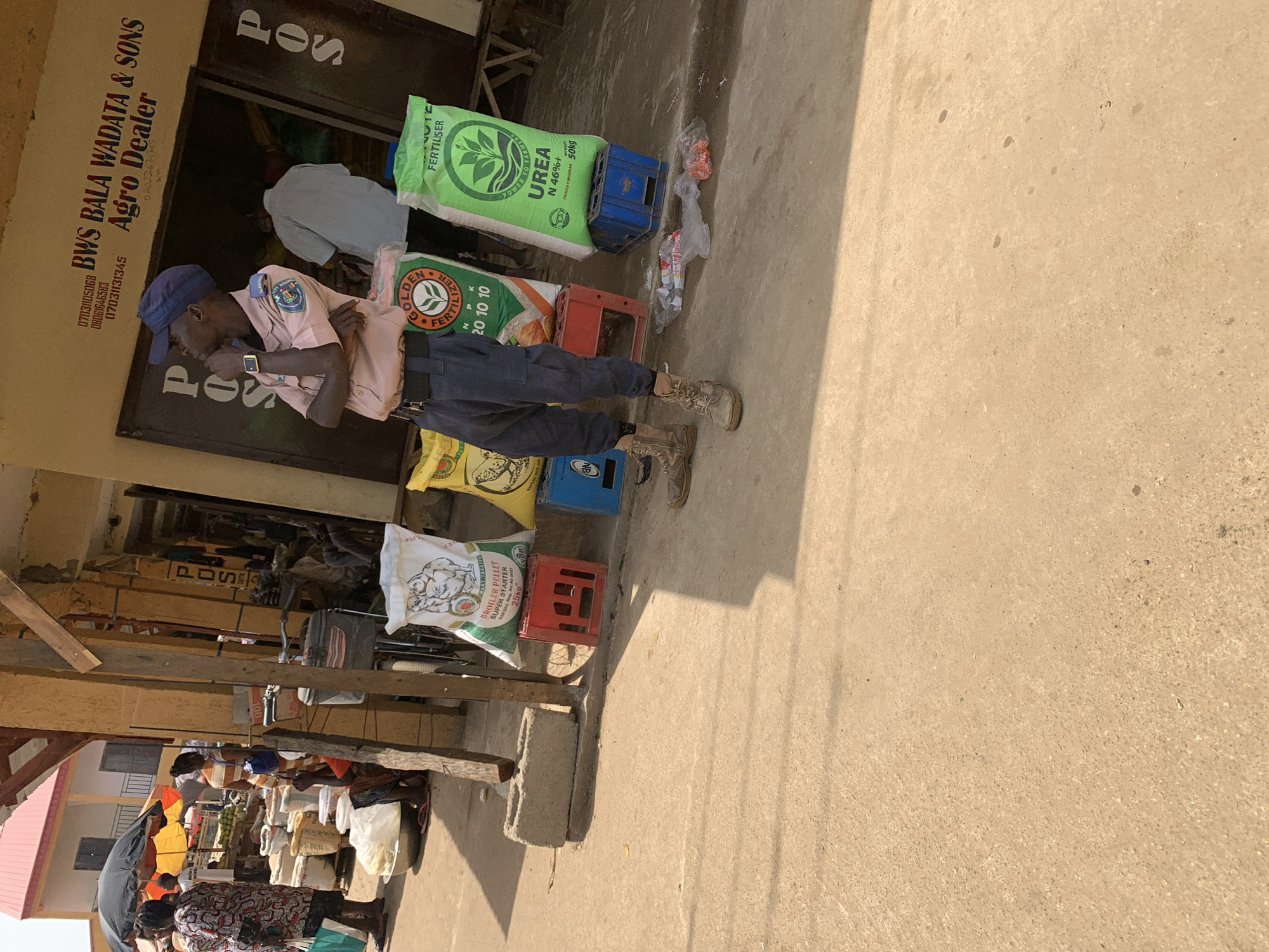 This media file captures a sunny street scene in Kafanchan B, Nigeria, featuring an agro-dealer shop and bustling market activity. The main subject is a man in a light pink uniform shirt, dark trousers, and a blue cap, wearing boots. He stands upright, his left arm folded across his chest, next to the entrance of a shop. A watch is visible on his left wrist.
The shop, identified by the signage "BWS BALA WADATA & SONS Agro Dealer," displays several large sacks of agricultural products outside. These include a green bag labeled "UREA N 46%+ FERTILISER PROUDLY NIGERIAN 50kg" and an orange/yellow bag with "GOLDEN FERTILIZER N P K 20 10 10." A partially visible white bag is labeled "BROILER PELLET SUPER STARTER 25kg." The bags are propped on red and blue plastic crates. The shop also features multiple "POS" (Point of Sale) signs, and contact numbers are prominently displayed on the wall: "0703105068," "08061646583," "07031131345," and "08022671705." Another person in a light blue shirt is visible inside the shop doorway.
The ground is concrete, with strong shadows indicating bright daylight, possibly morning or late afternoon. Some litter, including plastic bags, is scattered near the product sacks. Further down the street, to the left of the main shop, a vibrant market scene unfolds with several people gathered around stalls, engaged in daily commerce. Wooden structures and partial views of other buildings can be seen in the background.