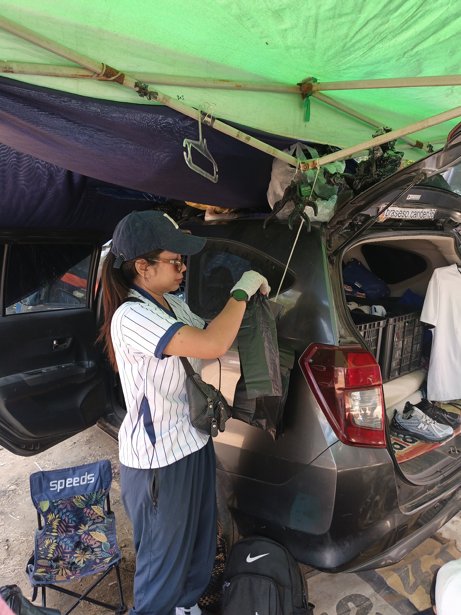 A woman wearing a blue baseball cap, sunglasses, a white and blue striped polo shirt, and dark blue pants stands next to the open trunk of a dark gray car. She is wearing light green gloves and holding a black plastic bag. The car is parked outdoors under a green tarp supported by metal poles, and a dark blue tarp is draped beneath it. In the background, the trunk is filled with various items, including a black crate containing hats, a white t-shirt, and other bags. A foldable chair with a floral pattern is positioned to the left of the car. The ground appears to be dirt and gravel. The overall scene suggests an outdoor market stall or a temporary setup for selling goods, possibly in Jatinom, Indonesia.