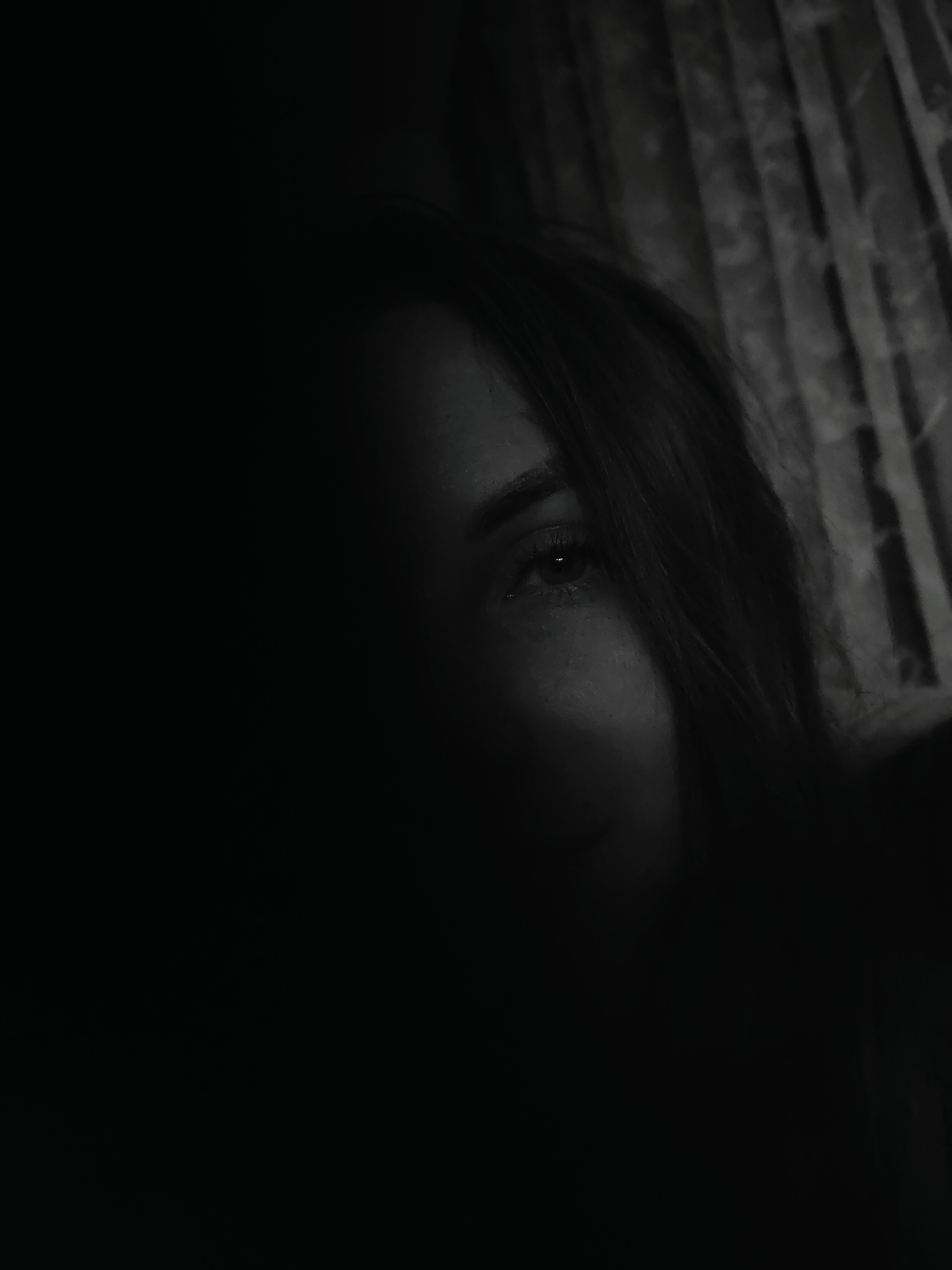 A monochromatic, close-up image depicts a female subject in an indoor, dimly lit environment. The primary focus is the upper right quadrant of her face, with her right eye, eyebrow, and a portion of her cheek visible. The left side of her face is obscured by deep shadow, and strands of dark hair partially cover her forehead and the left side of the frame. Her right eye is open and appears to gaze directly forward. The background, visible behind the subject's head to the right, consists of vertically oriented, textured elements, consistent with window curtains or blinds, rendered in varying shades of gray due to low illumination. No specific landmarks or unique architectural features are present to identify a precise location within Chaikovsky, Russia.