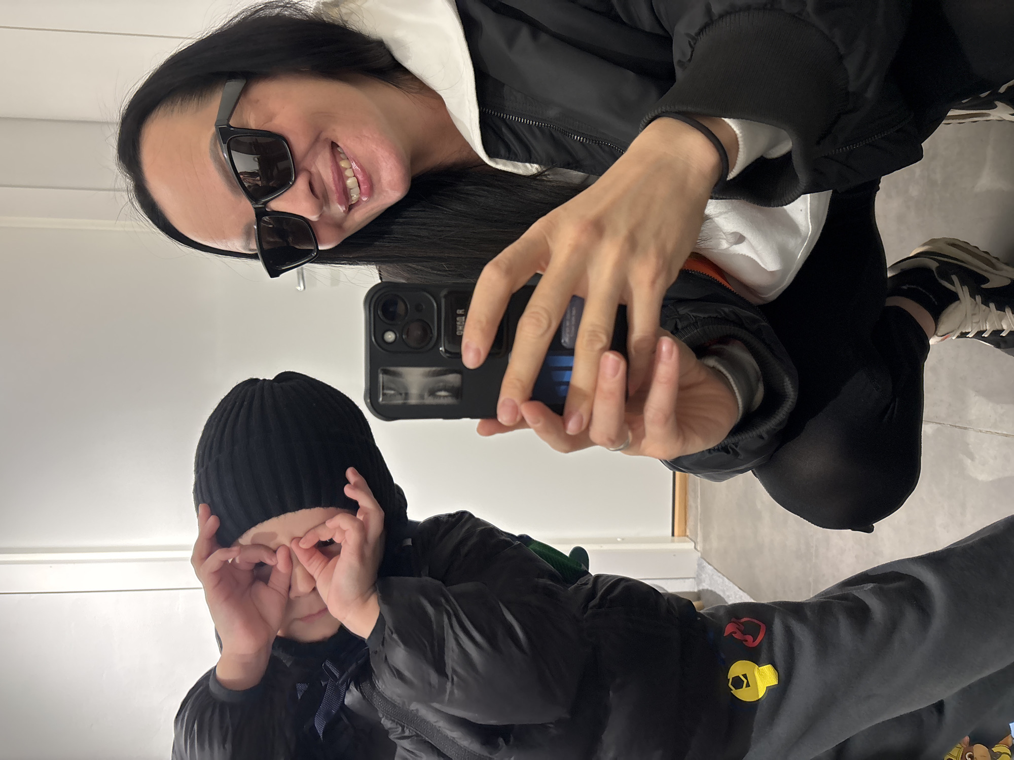 A woman in sunglasses and a black jacket, along with a child wearing a black beanie and jacket, are taking a selfie in what appears to be a dressing room or mirrored space. The woman is holding a smartphone, with her reflection visible on the screen. The child is making binoculars with their hands over their eyes. A black and white sneaker is visible. The location appears to be a mirrored room in Chaikovsky, Russia.
