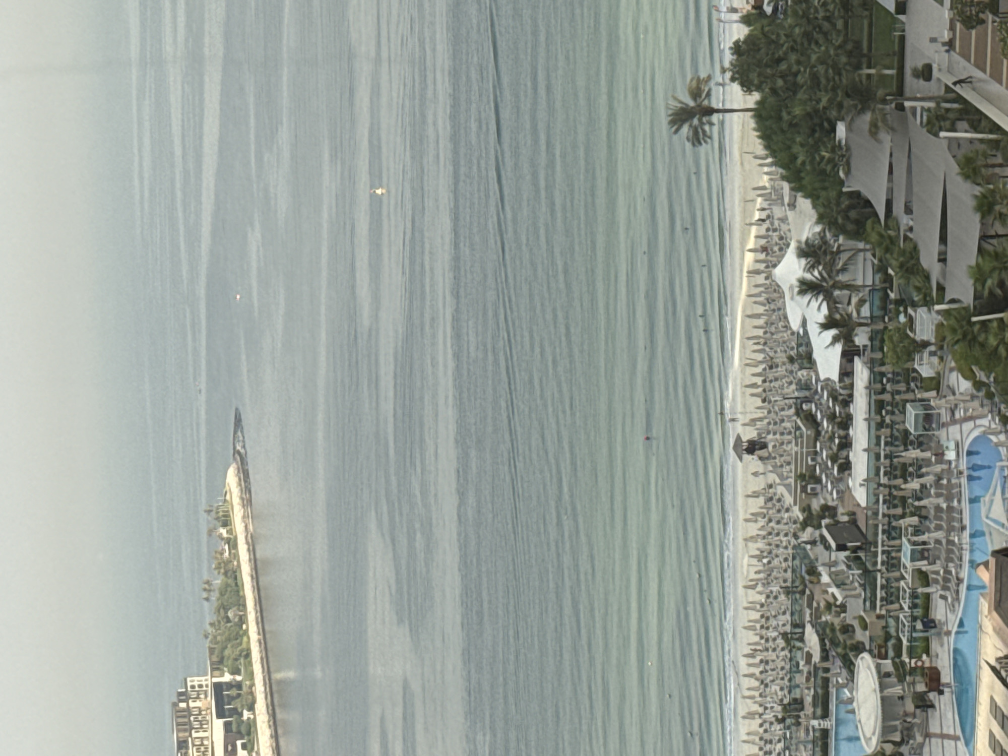 This image captures a panoramic view of a beach resort in Dubai, United Arab Emirates. In the foreground, several swimming pools and lounge areas are visible, adorned with numerous umbrellas and seating arrangements. Beyond the resort, a white sandy beach stretches along the coastline, dotted with more umbrellas. The turquoise waters of the Arabian Gulf extend to the horizon, with small waves rippling towards the shore. A modern hotel complex is situated on a man-made peninsula in the distance, overlooking the sea. Several people are seen enjoying the water and the beach, while a few boats are dotted across the ocean. The sky is hazy, characteristic of Dubai's climate.