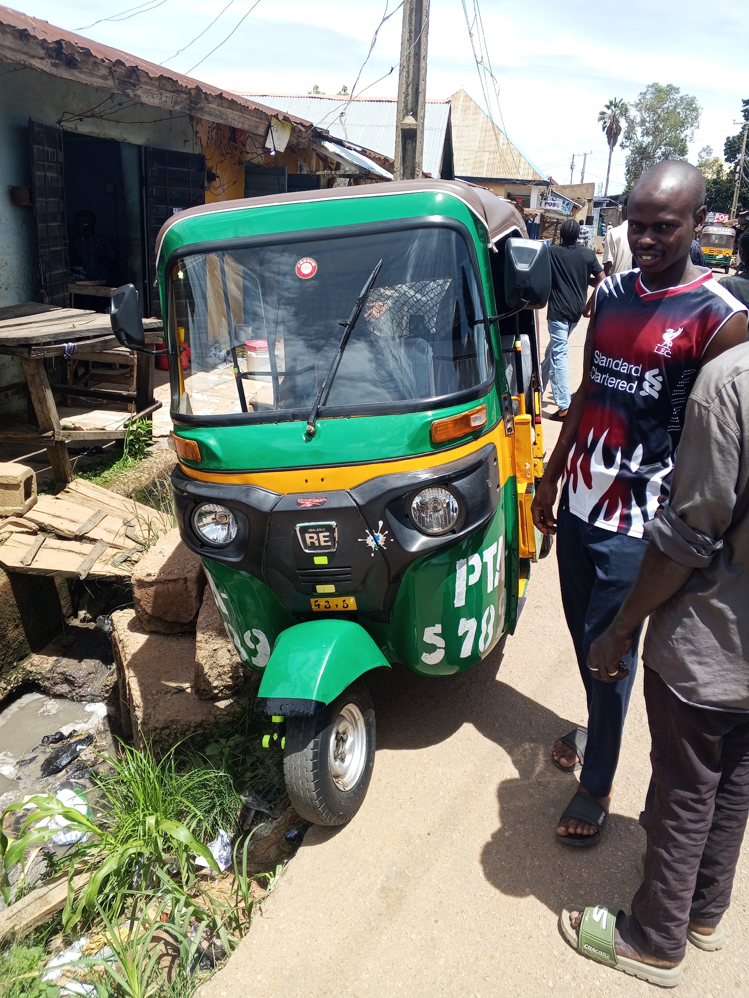 The image depicts a bright, sunny street scene in an unknown urban or semi-urban location. In the center, a three-wheeled auto-rickshaw (tuk-tuk) is parked, colored predominantly green with yellow accents. The vehicle displays "BAJAJ RE" branding on its front panel, along with the numbers "PTA 578" (partially obscured) and "43.5" in white lettering. A single headlight is visible on its left side, and a red circular sticker is affixed to the windshield.
To the right of the tuk-tuk stands a dark-skinned man with a bald head and light facial hair, wearing a red, black, and white sleeveless Liverpool FC jersey featuring a white flame-like design. He looks directly at the viewer with a slight smile. He wears dark blue trousers and black slide sandals. Partially visible next to him, to the right, is another dark-skinned man from the chest down, wearing a light brown long-sleeved shirt and dark trousers, with olive green slide sandals bearing the word "CHACRAS."
To the left of the auto-rickshaw, a narrow, open ditch or gutter runs along the roadside, containing stagnant water and various items of refuse, including plastic bags and bottles. Green grass and weeds grow along the edge of this ditch. Beyond it, a dilapidated single-story building with a corrugated metal roof has an open doorway revealing a dark interior. Makeshift wooden planks and structures are positioned outside this building.
In the background, the street continues with several other blurred figures walking or standing. Further buildings with corrugated roofs are visible, along with utility poles, overhead wires, and a distinct palm tree on the right side. The scene is illuminated by strong, direct sunlight, casting clear shadows on the paved ground.