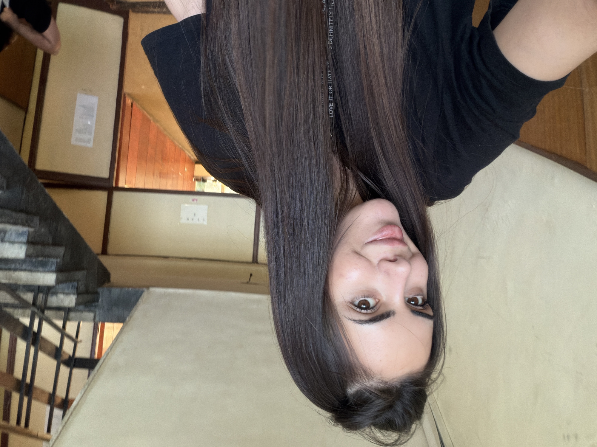 A young woman with long brown hair, wearing a black t-shirt, is pictured indoors in a corridor or stairwell. The camera angle is unusual, suggesting she is holding the camera above her head, resulting in an upside-down perspective. In the background, there is a staircase with a black railing. There are light-colored walls with what appears to be some type of wooden paneling and signage on the walls. Based on architectural characteristics, it's possible this is a residential apartment block built in the Socialist era, which is common in Sofia, Bulgaria. Without additional context or features, the precise location is indeterminate.