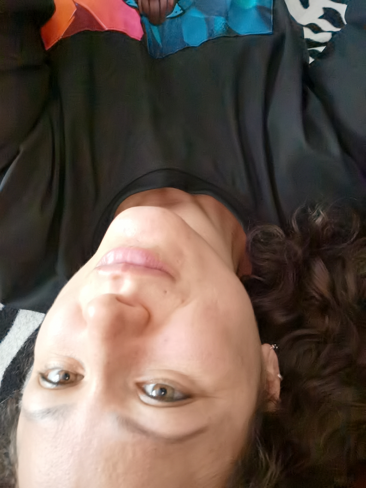 The image is an upside-down selfie of a person with curly brown hair. The person is wearing a black shirt with a colorful, abstract pattern on the upper chest area. The background appears to be a striped surface. The lighting suggests an indoor setting. There is no discernible information about the location in Chaikovsky, Russia.