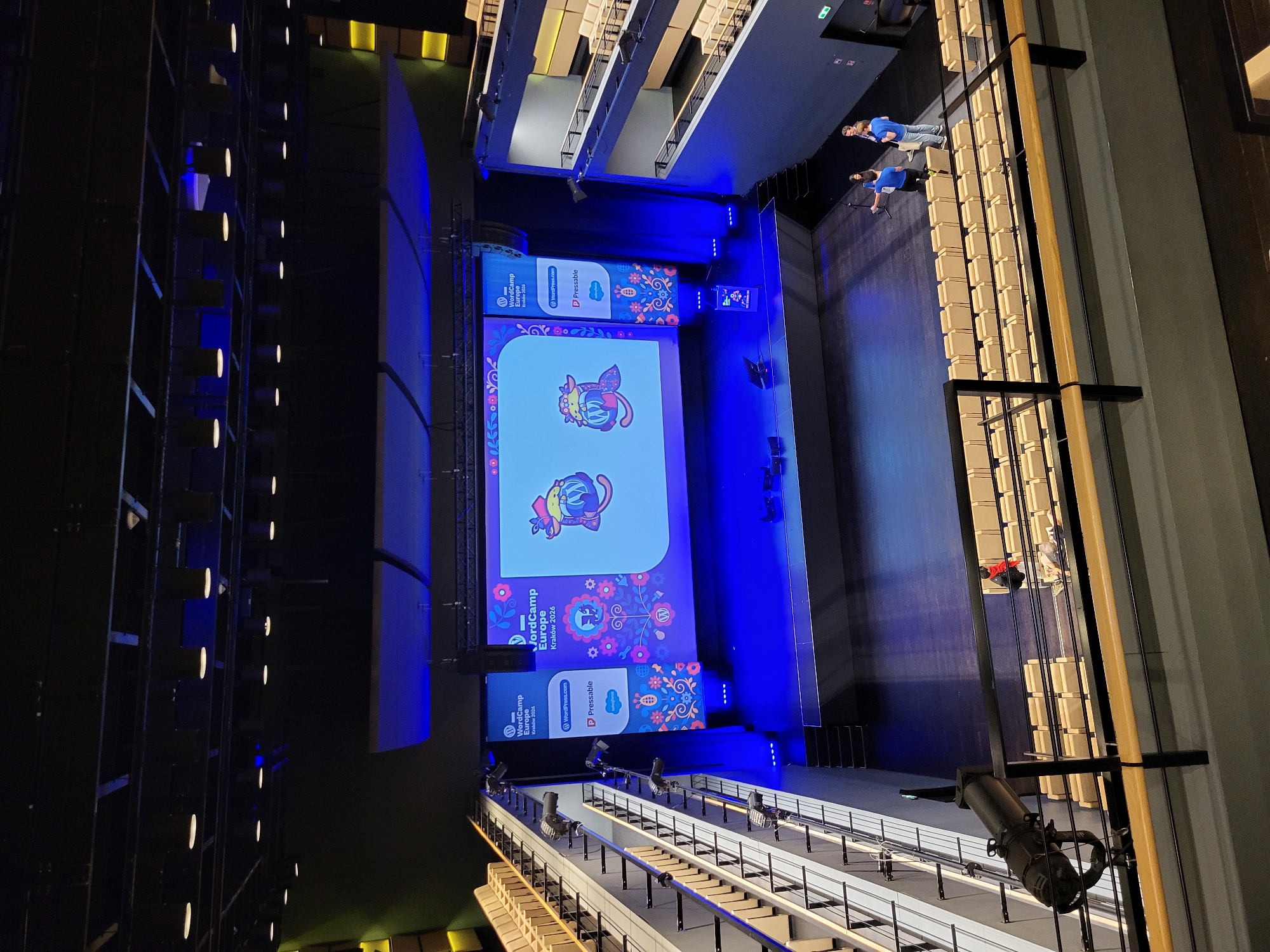 A high-angle photograph captures a wide view of an empty auditorium or large conference hall, likely in the city of Unknown, Unknown. The perspective is from an upper-level balcony, looking down towards a stage.
The stage, centrally located at the front, features a large, rectangular LED screen displaying "WordCamp Europe Kraków 2026" with the WordPress logo. The screen's background includes colorful, stylized folk-art patterns on the left and right edges, flanking two cartoonish, mythical animal figures (possibly a fox or badger-like creature with decorative elements). Two narrower, vertical banners or screens stand on either side of the main screen, repeating "WordCamp Europe Kraków 2026" and displaying logos for WordPress.com, Pressable, and Salesforce. The stage area is illuminated with a dominant blue light, casting a glow on the dark stage floor. A small dark table and two dark chairs are positioned on the stage, in front of the main screen.
Numerous light-colored, padded chairs are arranged in rows on the main floor directly in front of the stage, appearing empty. Additional levels of tiered seating or balconies, also with light-colored, empty chairs, line both the left and right walls of the auditorium. The seating on the left appears to extend upwards in steep tiers. The right side features two distinct balcony levels.
Three individuals are visible on the floor level, in front of the stage, towards the right side of the image. Two individuals wear blue t-shirts; one stands facing right, and the other kneels, facing the stage area. A third individual, positioned between them, appears to be observing or interacting with the kneeling person. Their actions suggest preparation or setup activity, possibly related to audio-visual equipment, as one individual appears to be holding a device.
The auditorium's ceiling is dark with exposed rigging and numerous black stage lights mounted. Additional spotlights are positioned along the balcony railings, including a large black spotlight in the immediate foreground, aimed towards the stage. The walls are dark, with sections appearing to have acoustic paneling.