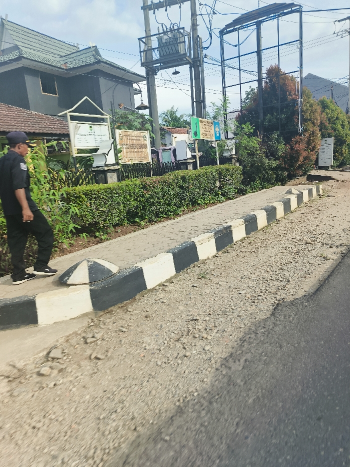 The image shows a street scene in Sukatani, Indonesia. A man in a black uniform and a black cap is walking on a sidewalk. He is wearing a black belt and black shoes. The sidewalk is paved with gray concrete and has a black and white curb. The sidewalk is bordered by a row of green bushes. There is a power pole with an electrical transformer on it to the left of the image. There is a tall structure to the right of the image that appears to be a billboard. There is a green sign on the wall of a building in the distance. The street surface is rough and made of gravel and asphalt. The background of the scene is a row of trees. The sky is cloudy and there is a mountain visible in the background.