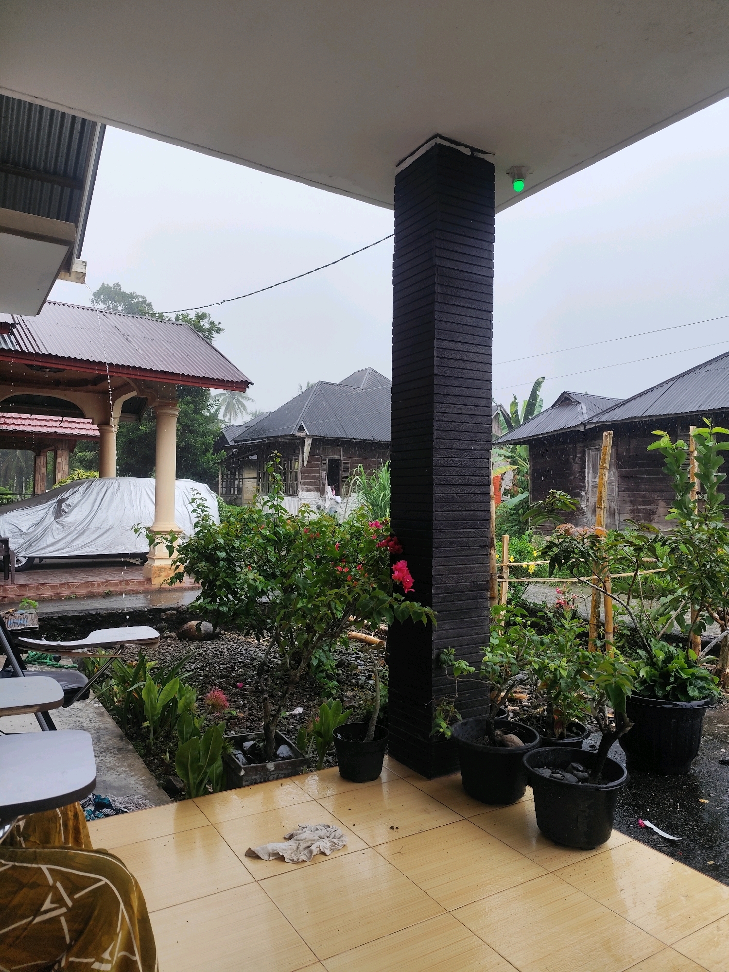 The image shows a covered car parked under a covered patio area during a rain shower. The patio has a tiled floor that is wet from the rain, and a black, brick-patterned pillar supports the overhang. Several potted plants, including some with pink flowers, are arranged near the pillar and in the foreground. Beyond the patio, there are traditional wooden houses with dark metal roofs, and lush green vegetation. The sky is overcast and the rain is visible as streaks in the air and on surfaces. In the background, palm trees are visible. A green light is illuminated on the ceiling of the patio near the pillar. The scene appears to be a residential area in Bonjol, Indonesia.