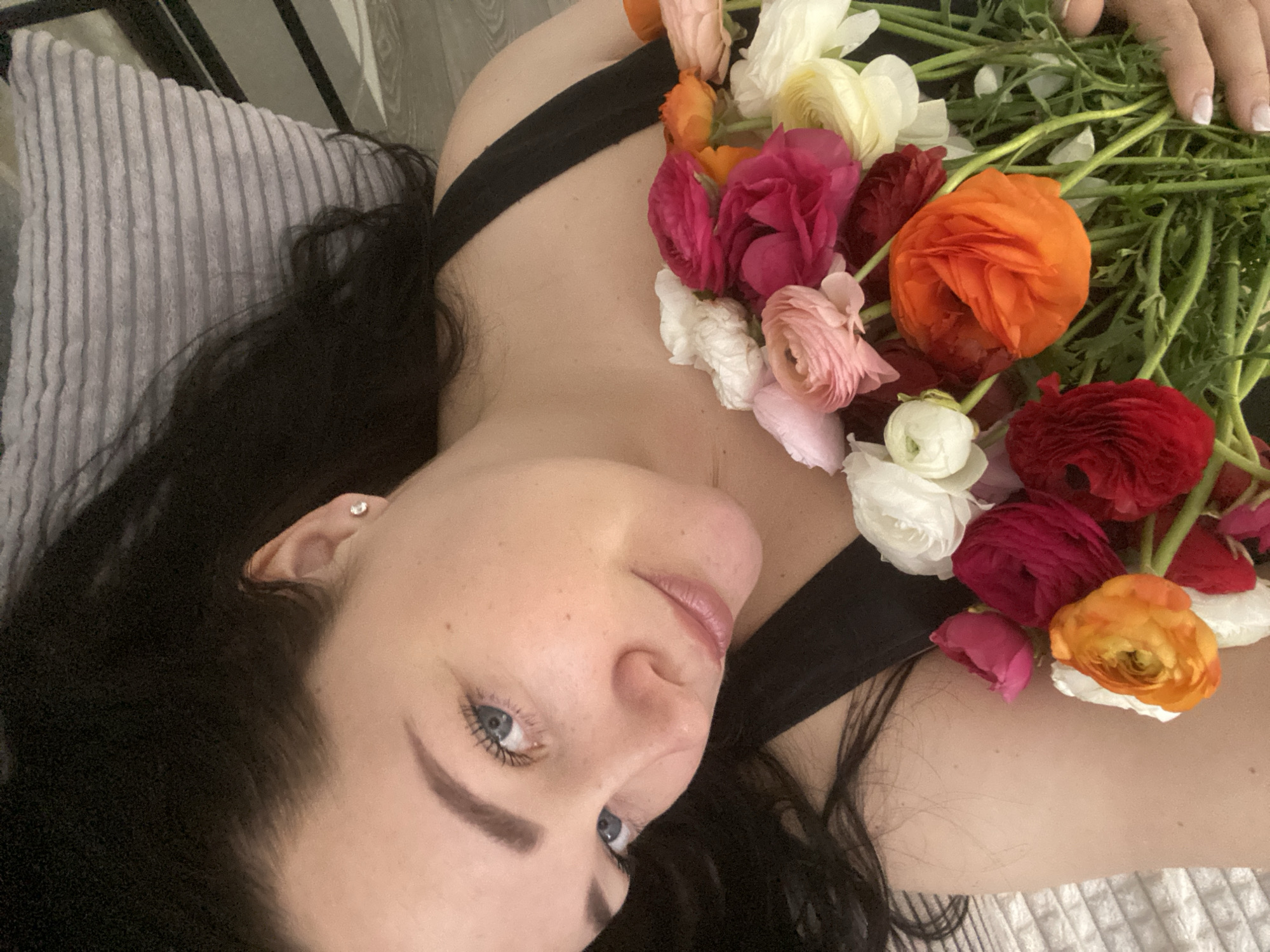 A woman with dark hair and fair skin is lying on a pillow, holding a bouquet of ranunculus flowers. The flowers are various colors including white, pink, orange, and red. The woman is wearing a black top. She has blue eyes and is looking up toward the camera. An earring is visible in her left ear. The background is a striped pillow and part of a metal bed frame. The image was not taken in Sochi, Russia.