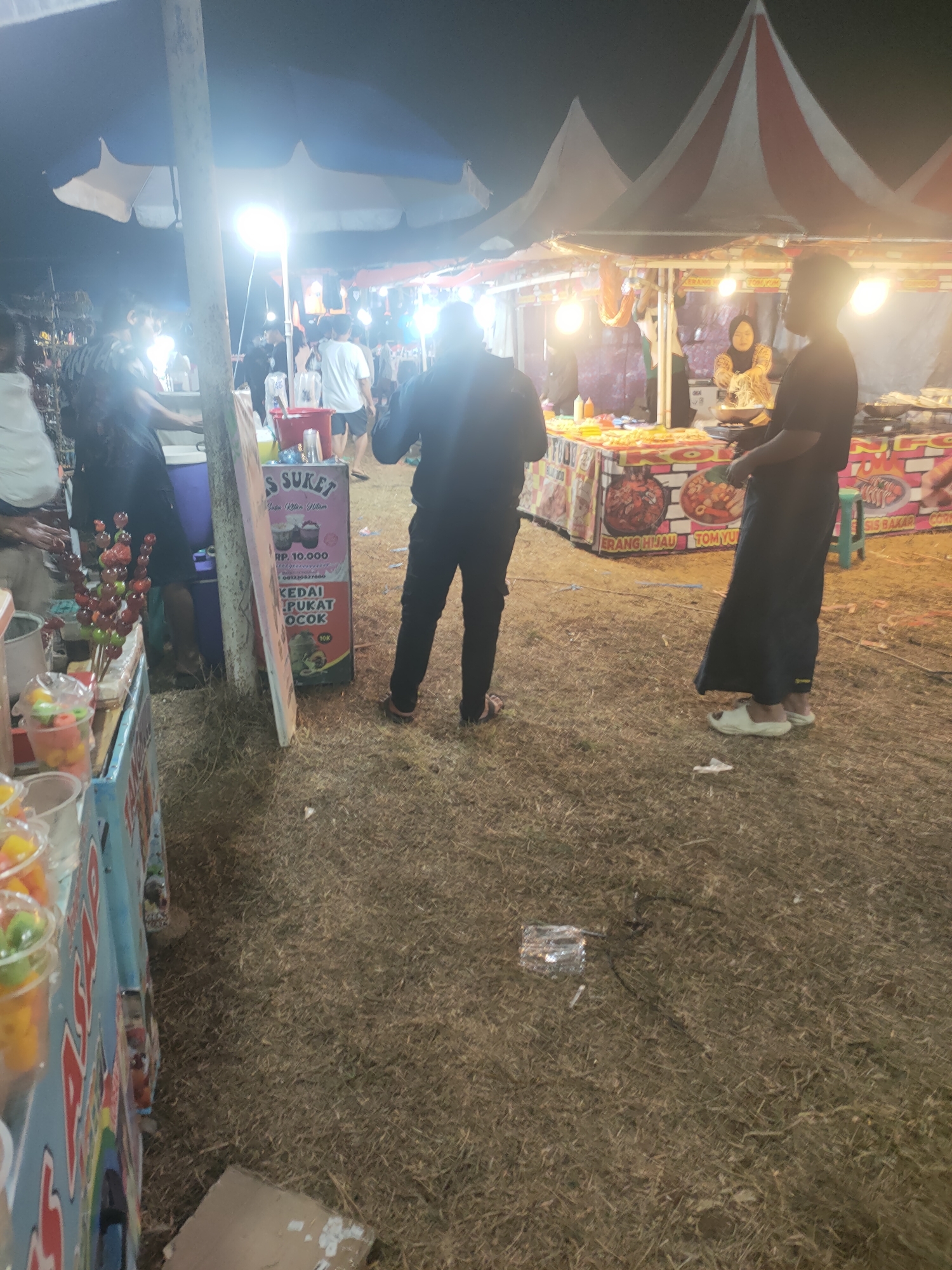 The image depicts an outdoor nighttime scene, likely a market or fair, in Subang, Indonesia. Several food stalls are visible, adorned with colorful tents and illuminated by string lights and spotlights. In the foreground, a vendor displays various colorful candied fruits on sticks, alongside cups of fruit salad. A sign with Indonesian text and prices is also prominently displayed, suggesting transactions are taking place.
Several individuals are present in the scene. Two people, one in a dark outfit and another in a black abaya, stand near the foreground stalls. Further back, other people are seen interacting at the food stalls, with a woman in a hijab visible behind a counter. The ground is covered with dry grass and scattered debris. The overall atmosphere suggests a bustling evening event.