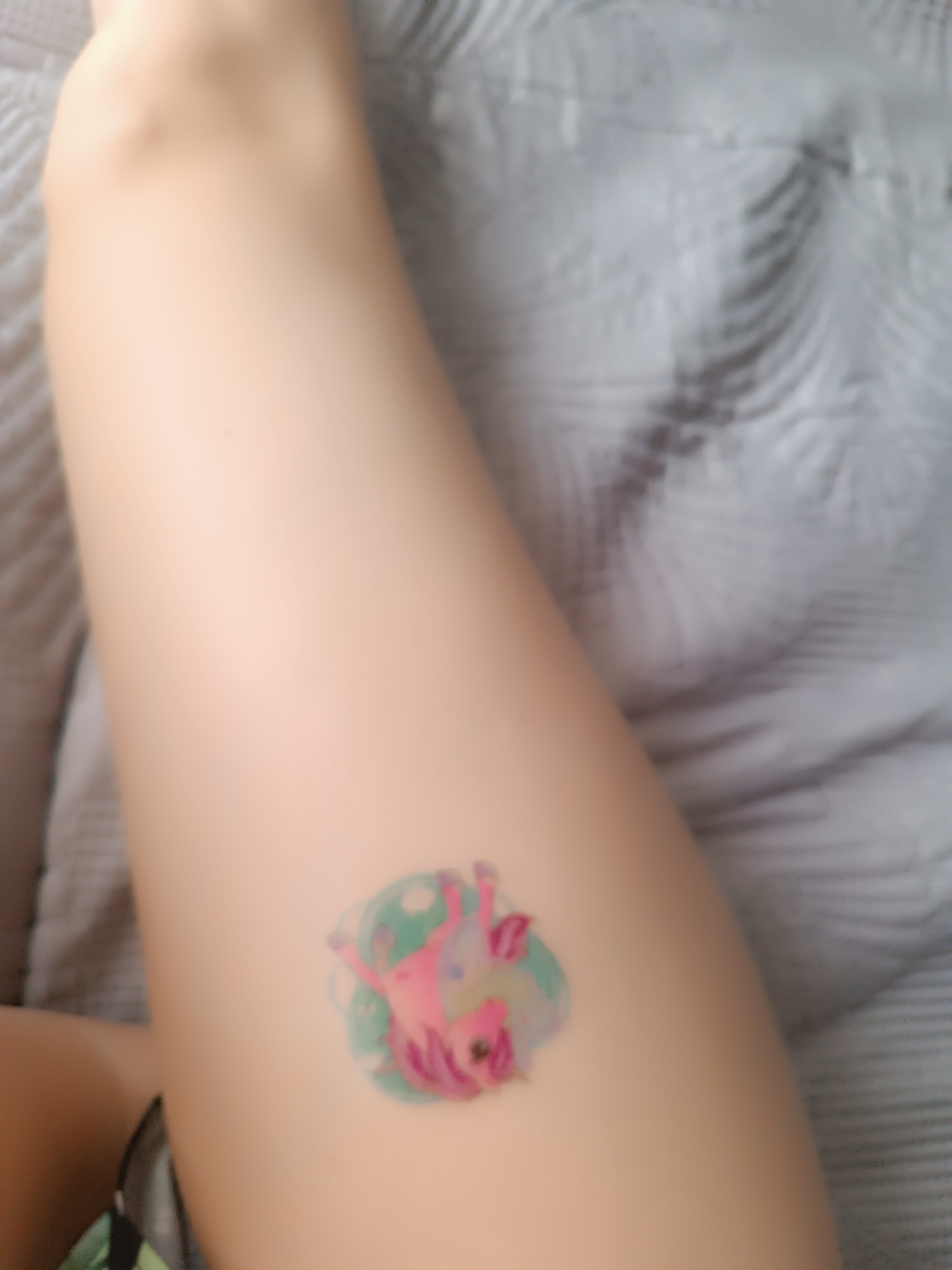 A blurred image shows a light-skinned thigh with a temporary tattoo of a pink unicorn surrounded by a teal, swirled border. The leg is set against a light gray, textured blanket. The location is unknown.