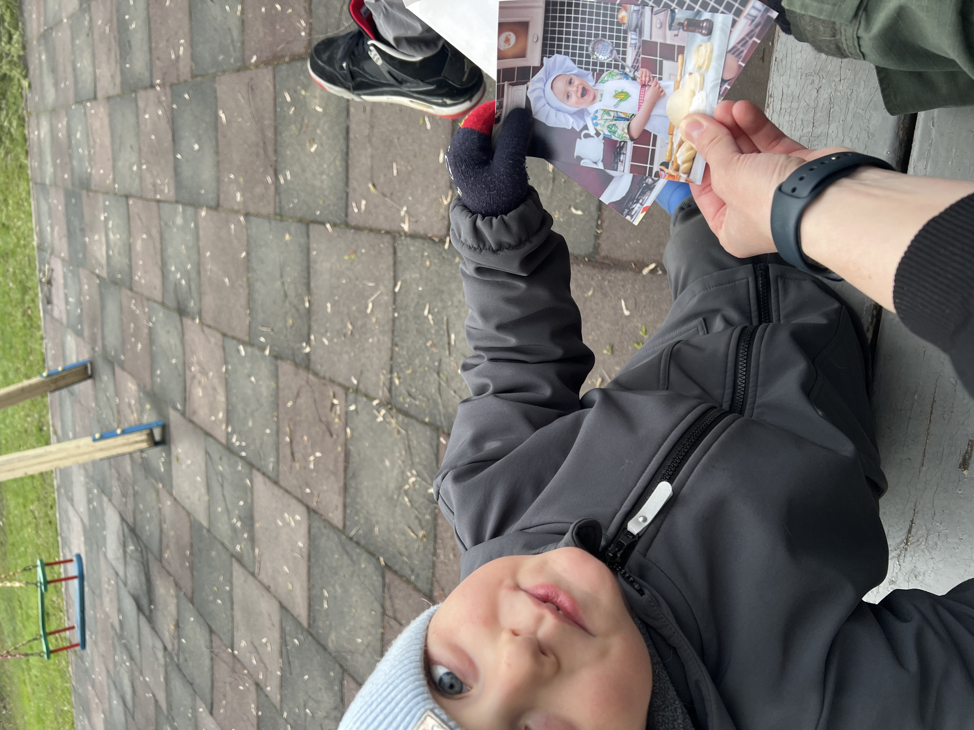 An outdoor scene captures a child and an adult interacting with a photograph, likely in a park or playground in Mytishchi, Russia. The child, seen from a low angle, looks directly at the camera with wide blue eyes and a slight pout. They wear a grey softshell jacket with a prominent black zipper and a light blue beanie. Their left hand, covered by a black glove with red fingertips, is raised towards the photograph.
An adult's hands hold a printed photo firmly. One hand, wearing a black sports wristband, supports the bottom of the photo, while the other hand, partially obscured, seems to be presenting it. The photograph depicts a smiling baby dressed as a chef, wearing a white chef's hat and a patterned apron, holding a spoon and standing in a kitchen-like setting with tiled walls and a countertop with baked goods.
In the upper right, part of an adult's leg and foot are visible, wearing a black sneaker with red accents and white sole detailing. The background features a paved ground made of interlocking grey and brown rectangular stones, speckled with small white debris, possibly petals or seeds. Patches of green grass are visible along the edges. To the left, wooden posts and blue/red elements suggest a swing set. Part of a dark, weathered wooden surface, possibly a bench, is visible on the right. The scene appears to take place during the day, likely in cooler weather given the warm clothing. No legible text is discernible on the child's beanie or jacket zipper pull.