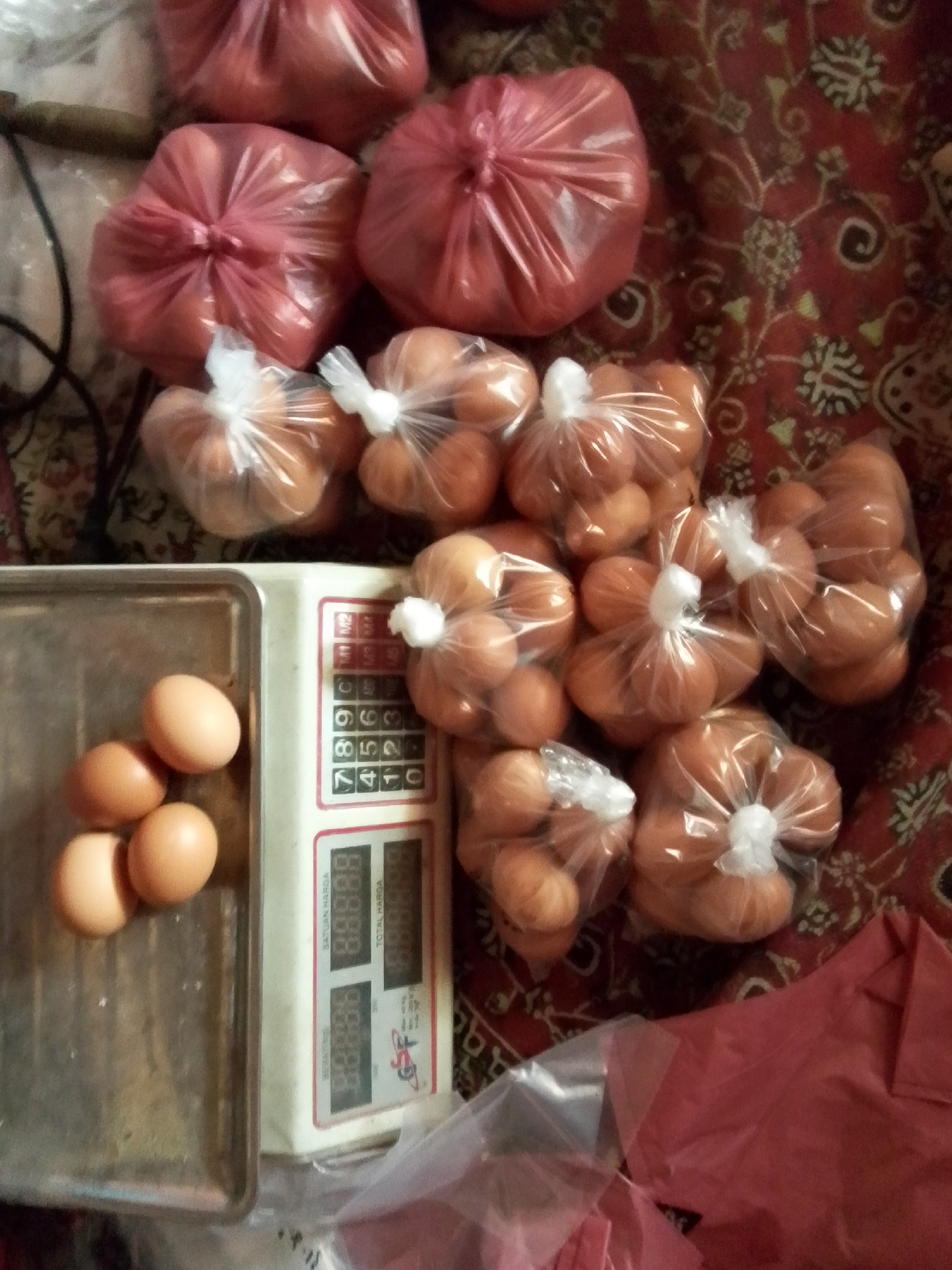 The image depicts a close-up, overhead view of a collection of eggs, some loose on a metal tray and others packaged in clear plastic bags tied with white ties. The eggs are light brown in color. Several larger, opaque red plastic bags are also visible, some containing what appear to be more eggs or a similar rounded product. A digital scale with a red and white casing is positioned to the right of the metal tray, displaying numerical readings. The background is a patterned red fabric, likely a tablecloth or rug. The scene appears to be indoors, given the lighting and arrangement of objects. No individuals are present in the frame. The location is identified as Bulurejo, Indonesia.