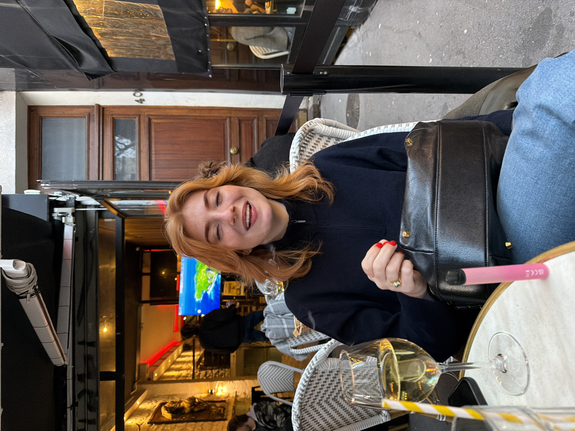 A woman with red hair is seated at an outdoor cafe in Paris, France. She is wearing a navy blue sweater and jeans and is smiling broadly. A black purse rests against her left side. A wine glass sits on the table in front of her, along with a pink disposable vape pen. The cafe has woven chairs, and a black awning is overhead. The building across the street has a wooden door with the number "40" above it. In the background, there is an indoor area with a television showing a map. The indoor space is lit with red and orange lighting, and other patrons are visible.
