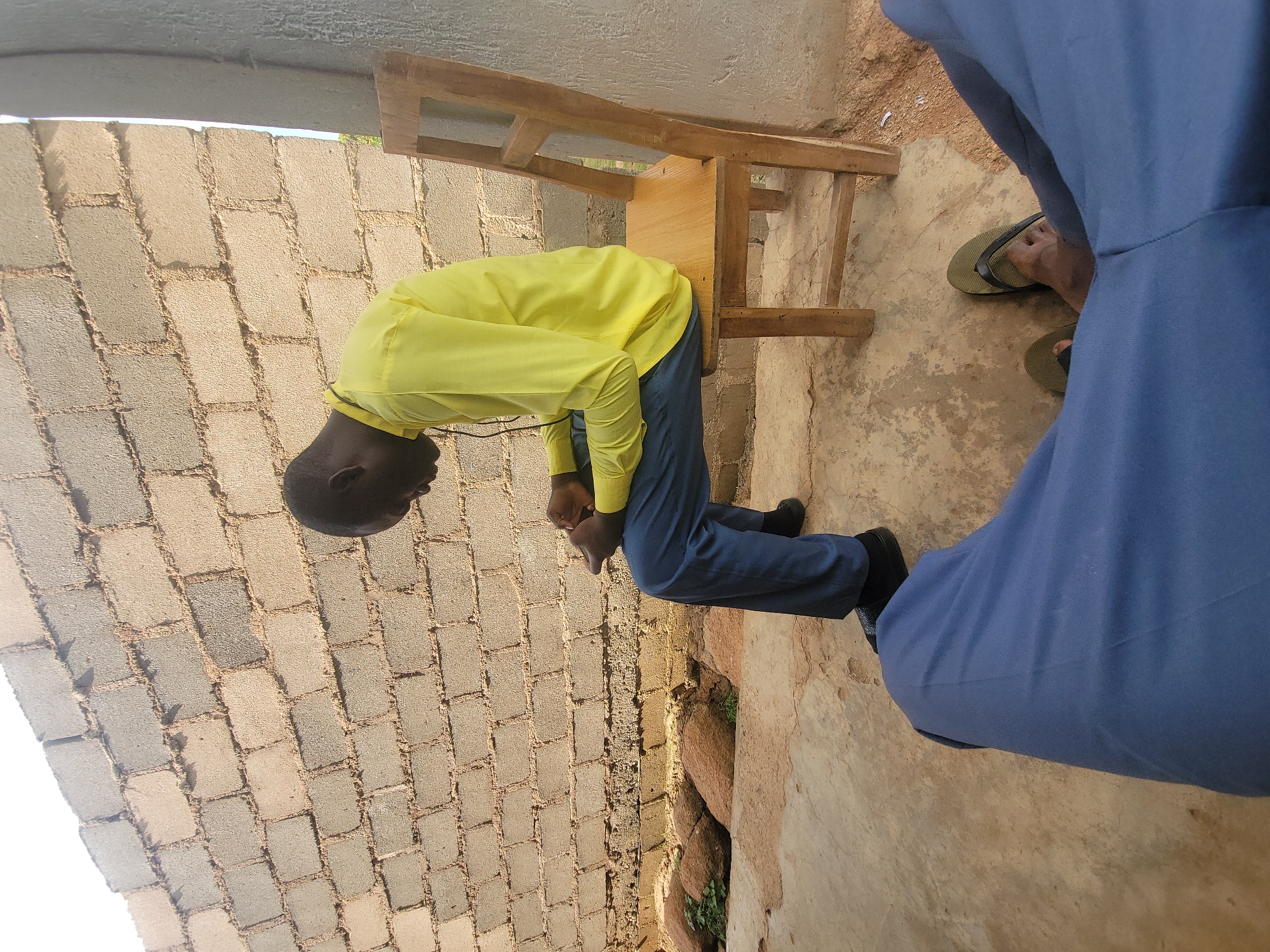 A man, appearing to be of African descent, sits facing right on a wooden chair. He is wearing a bright yellow long-sleeved shirt and dark blue trousers. His head is bowed, and he is looking down at something in his hands. The man is positioned outdoors, with a brick wall behind him and a concrete floor beneath him. To his right, the wooden chair he is sitting on is visible, and to his left, a person's legs wearing blue pants and green sandals are in the foreground. In the background, large rocks and some greenery are scattered near the base of the brick wall. The lighting suggests it is daytime. The scene is likely in an outdoor, possibly rural or semi-constructed area.